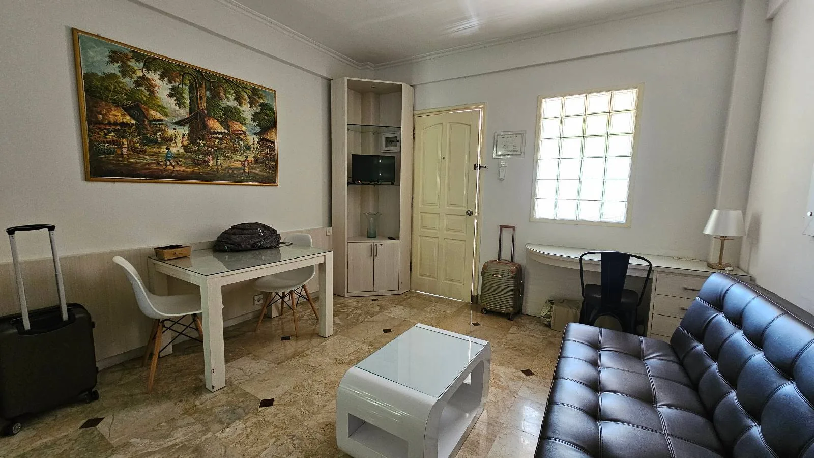 A small, tidy living room with a black sofa, a white coffee table, a desk with a chair, and a dining table with two chairs. There are two suitcases, a painting on the wall, and a window letting in light.