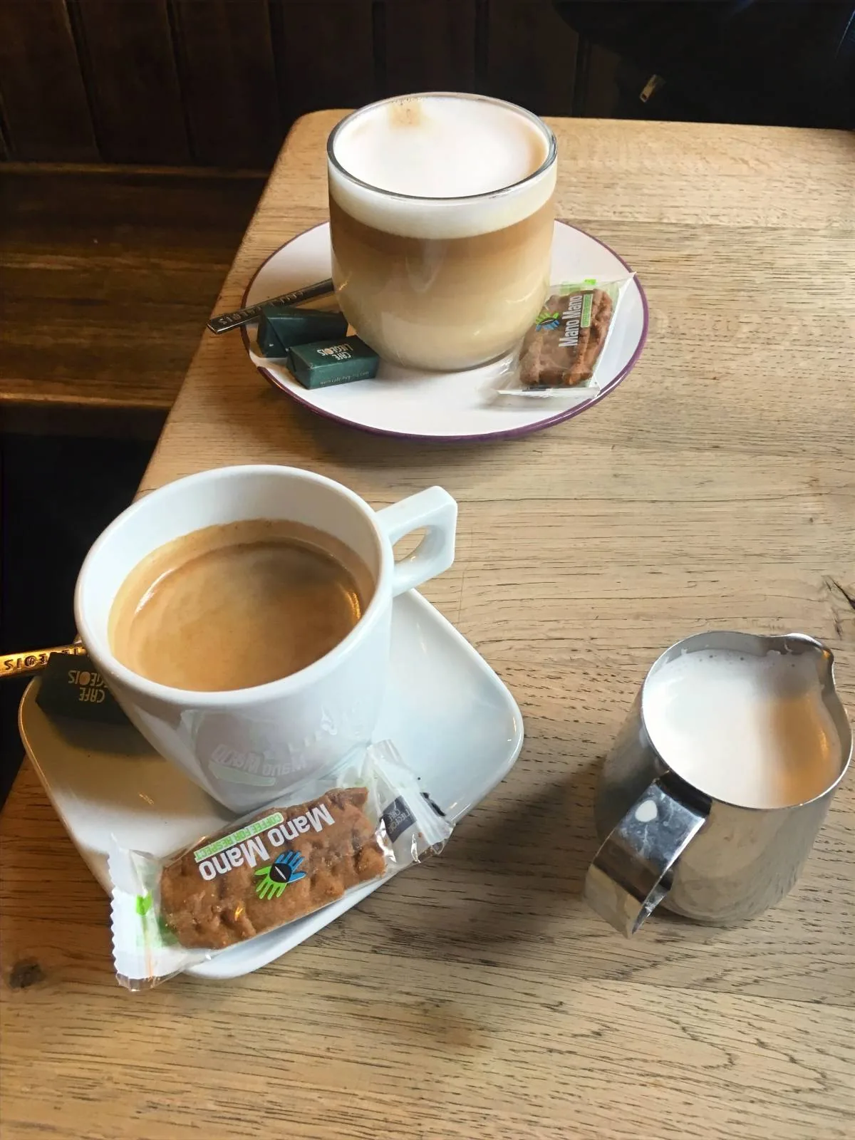 Two cups of coffee sit on a wooden table, each accompanied by a small wrapped biscuit. The cup in the foreground is white and paired with a small jug of milk. The background features a cappuccino with a saucer.