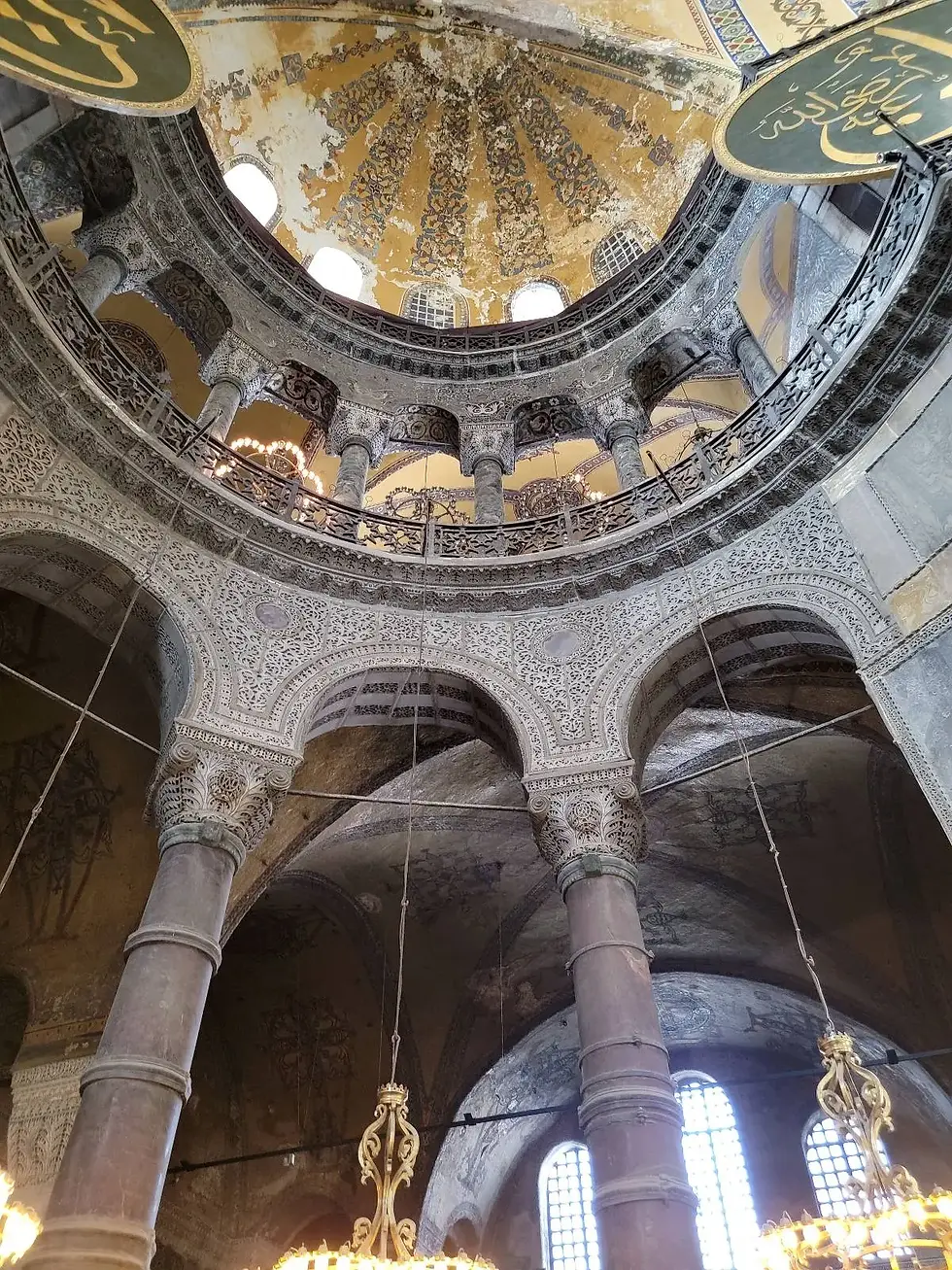 Interior of a grand domed building with ornate chandeliers and intricate mosaics on the ceiling. Natural light filters through arched windows.