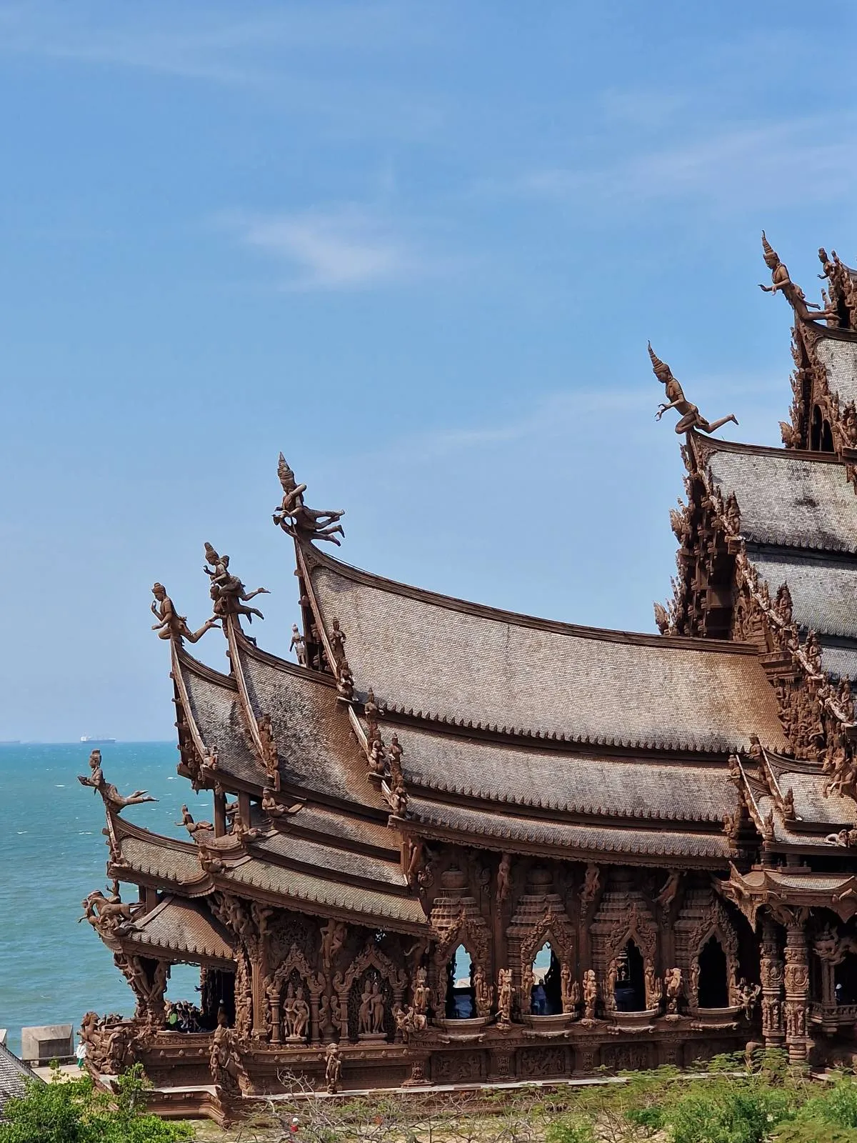 A detailed wooden temple with intricate carvings stands against a backdrop of a blue ocean and clear sky. The temple's ornate roof structures are prominently featured in the foreground.