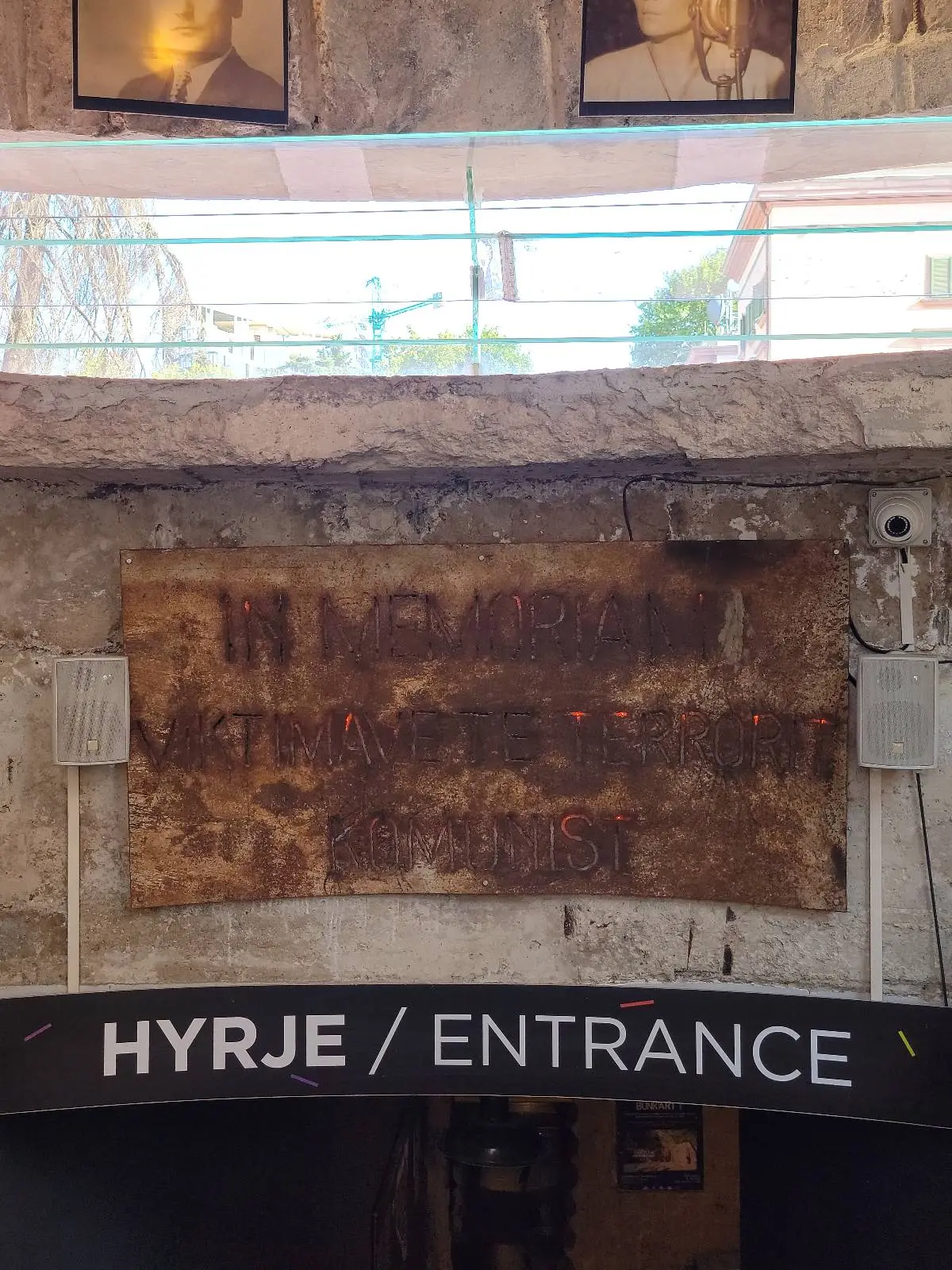 A sign reading "HYRJE / ENTRANCE" is displayed above a doorway. There is a large, textured brown panel or artwork above the sign. Sunlight filters in through a window above, casting light on the scene.