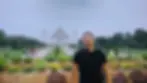 Smiling man in a black shirt poses in front of the Taj Mahal, with lush greenery and people walking nearby on a cloudy day.