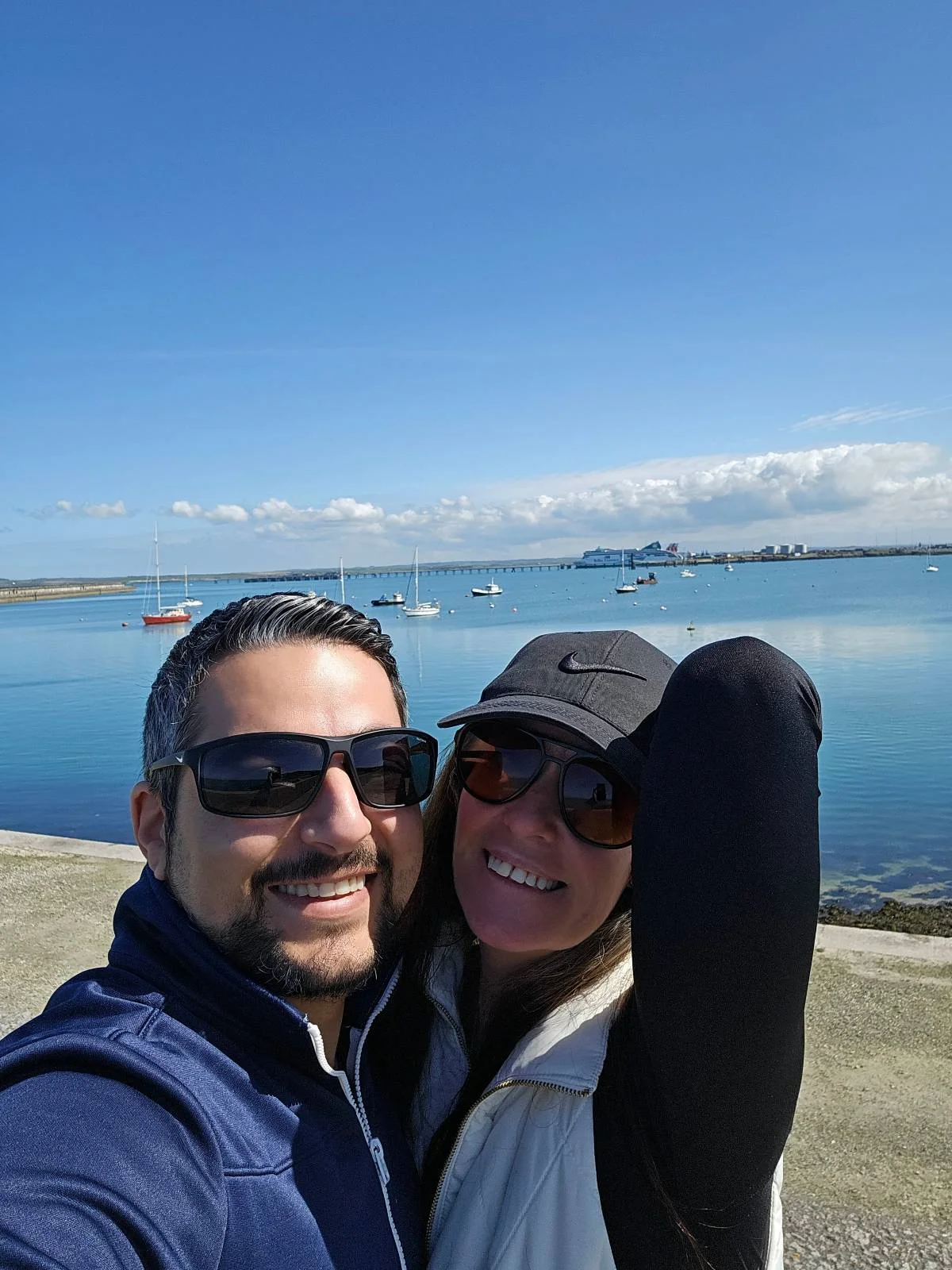 A couple wearing sunglasses smiling for a selfie with a serene sea and clear sky in the background.
