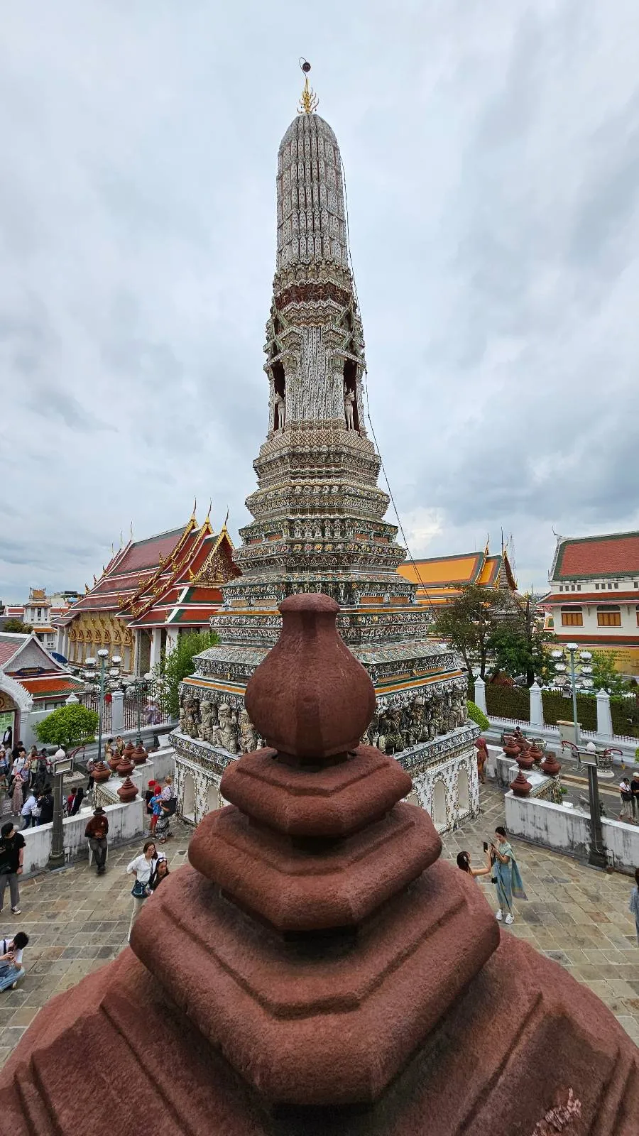 Tall, ornate spire with intricate details rises against a cloudy sky. In the background, traditional Thai temple buildings with colorful roofs and gold accents are visible. Visitors can be seen walking around the temple grounds.
