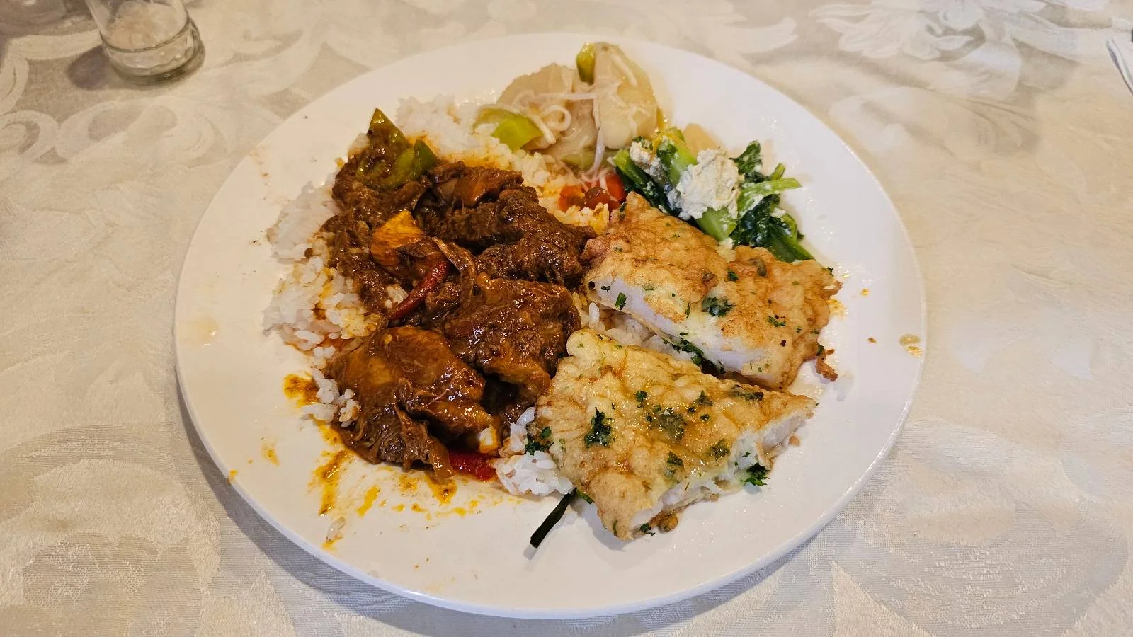 A plate with pieces of stewed meat, rice, herbs, and two breaded fish fillets on a white tablecloth.