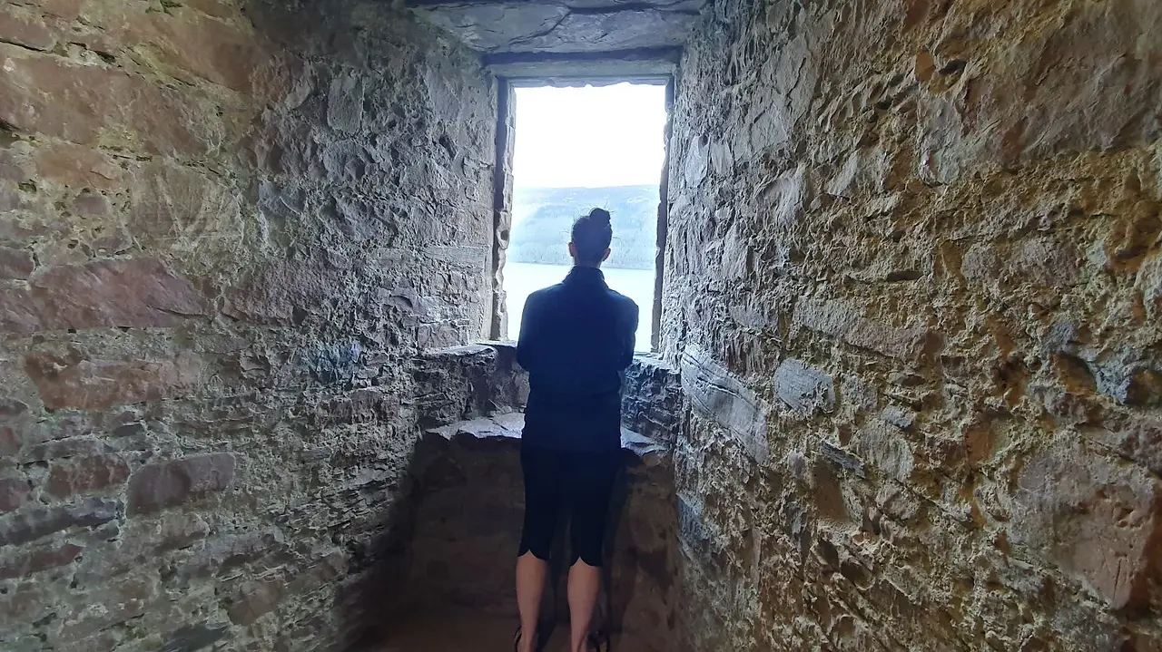 Person standing inside a stone room at Urquhart Castle, looking out through a window toward Loch Ness.