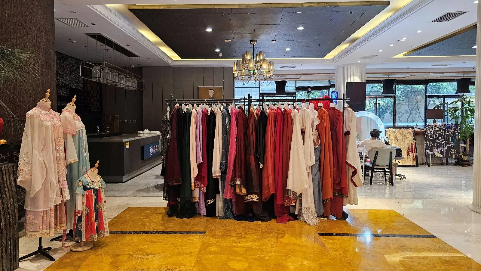 A clothing rack filled with colorful garments stands in the center of a well-lit, modern room with a chandelier, mannequins dressed in robes on the left, and a person working at a desk near large windows in the background.