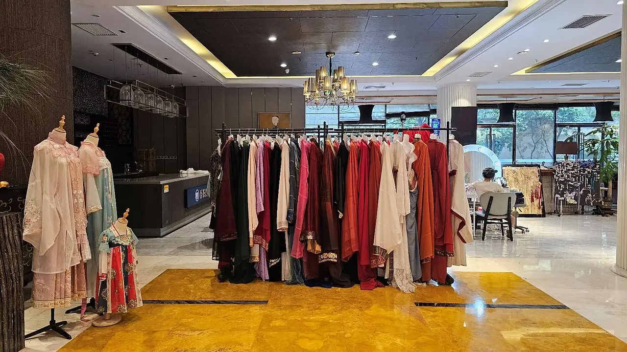 A clothing rack filled with colorful garments stands in the center of a well-lit, modern room with a chandelier, mannequins dressed in robes on the left, and a person working at a desk near large windows in the background.