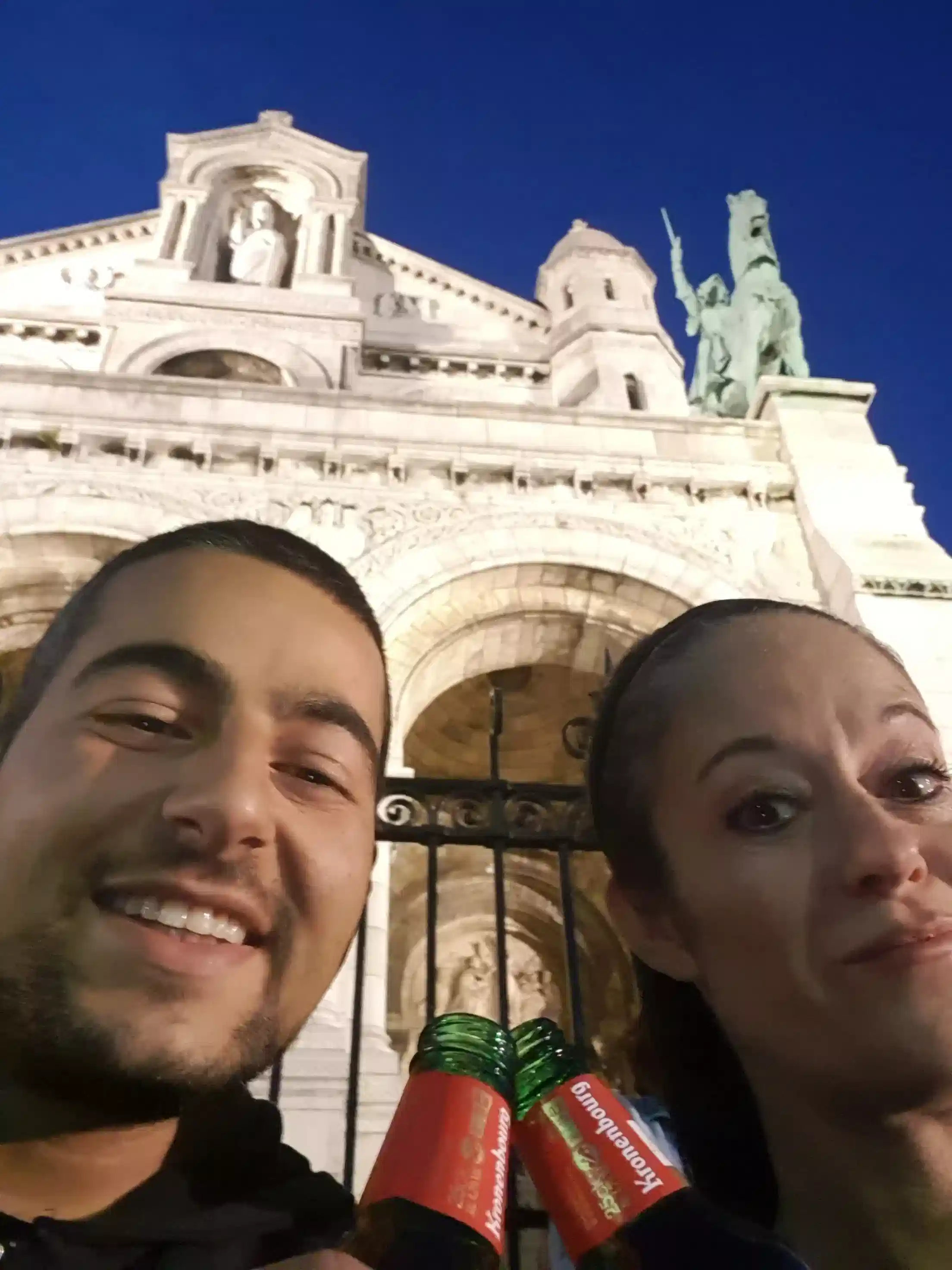 A man and woman smile and pose for a selfie in front of a grand, illuminated stone building with statues and arches at night.