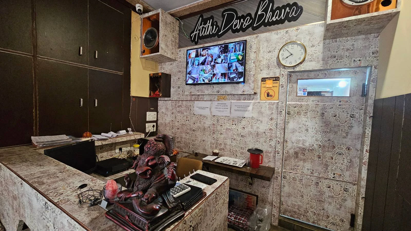 A counter with tiles featuring electronic equipment, a computer, and a backpack. Above, a flat-screen TV displays multiple security camera feeds. A clock and decorative speakers are mounted on the tiled wall, with "Jaipur Darbar Biryani" signage above.