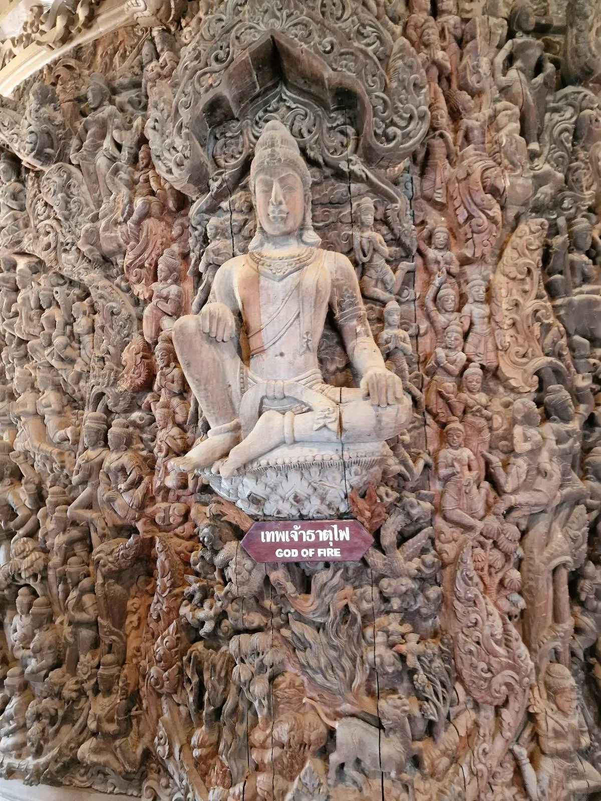 Intricately carved wooden sculpture depicting a seated figure surrounded by detailed patterns and designs on a large wall. The artwork includes a sign with decorative scripts beneath the main figure.