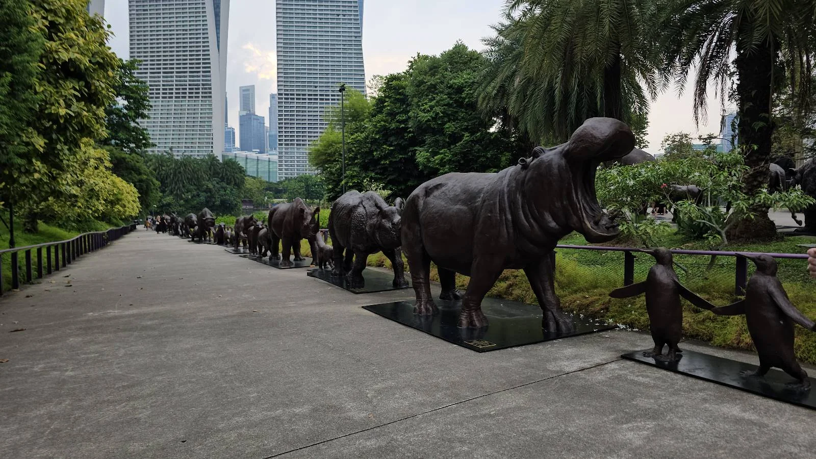 Pathway lined with bronze statues of elephants and other animals in a park setting, with tall buildings and greenery in the background. The sky is overcast.