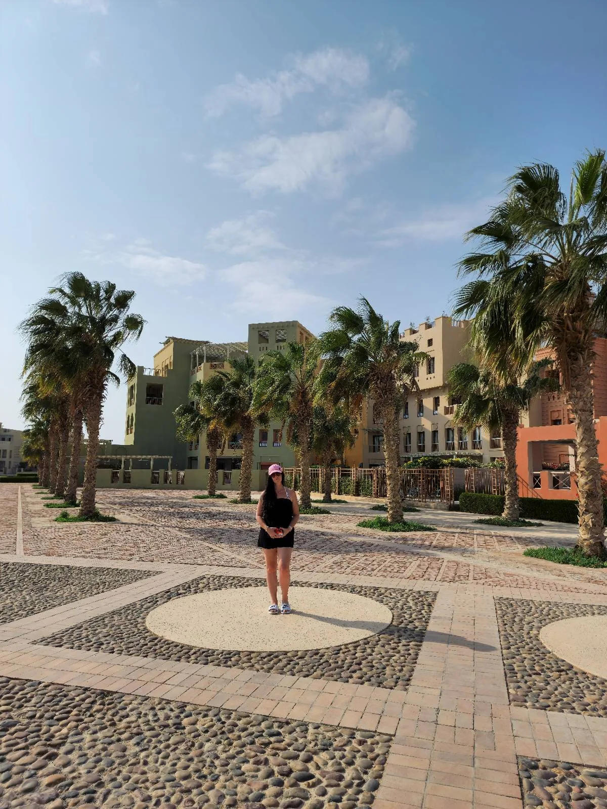 A person stands in a paved courtyard surrounded by palm trees and tall buildings under a clear blue sky.