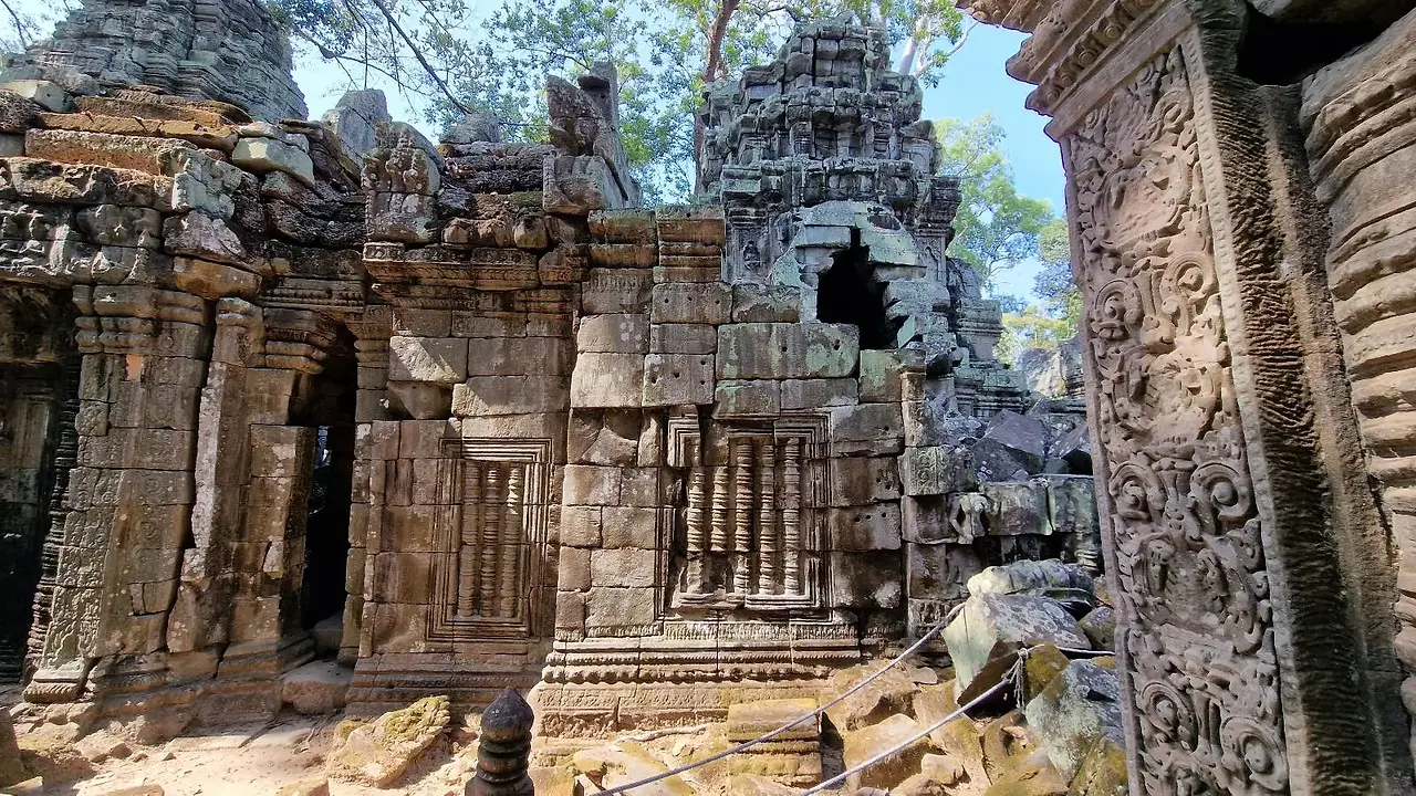 Close-up view of a crumbling ancient stone wall inside the ruins of Ta Prohm temple, with scattered rocks and debris below a large, jagged opening in the ceiling that reveals the blue sky and surrounding green trees.