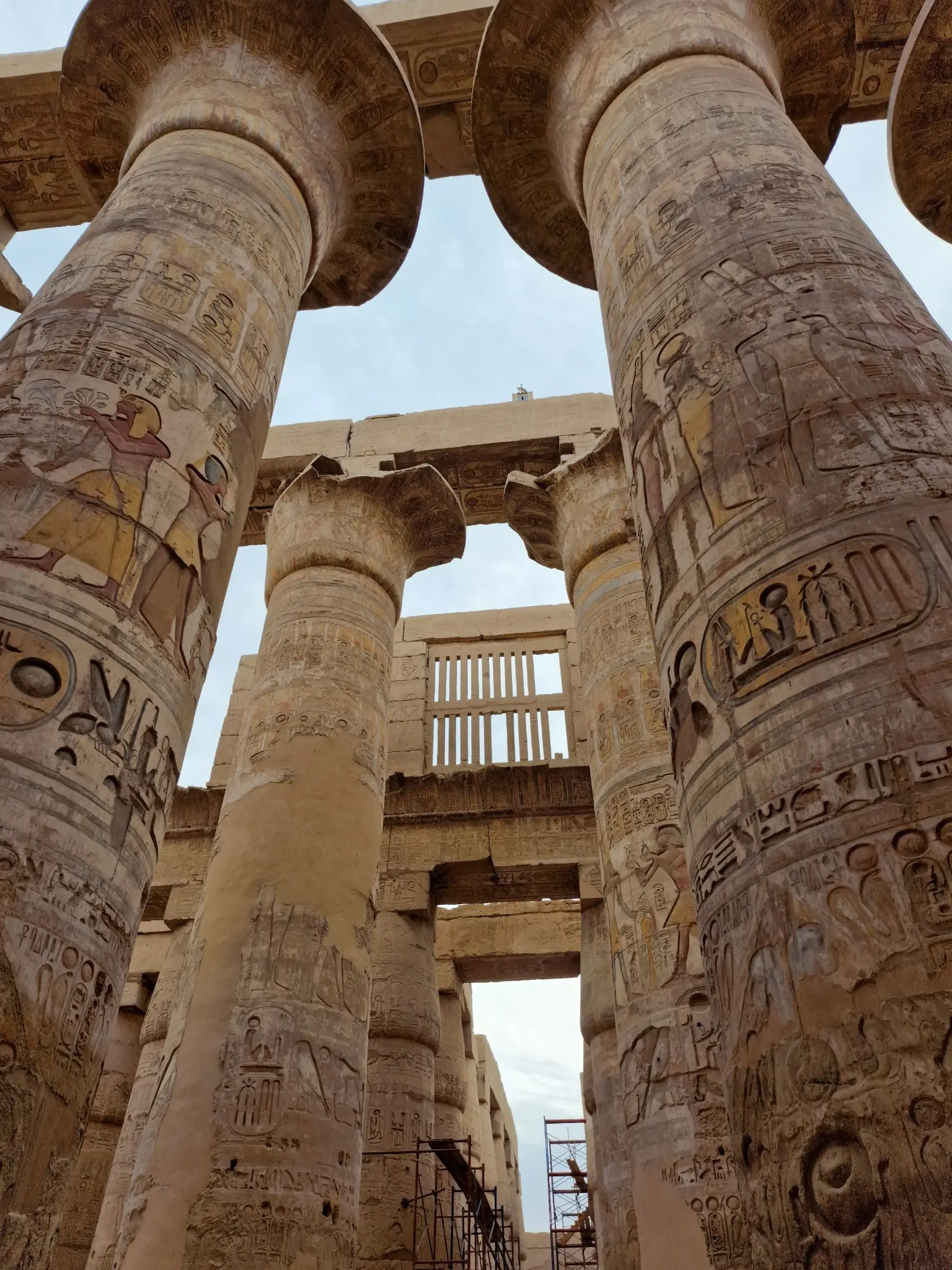 Ancient stone columns with hieroglyphics and carvings, part of Karnak Temple in Luxor, Egypt, are shown against a partly cloudy sky.