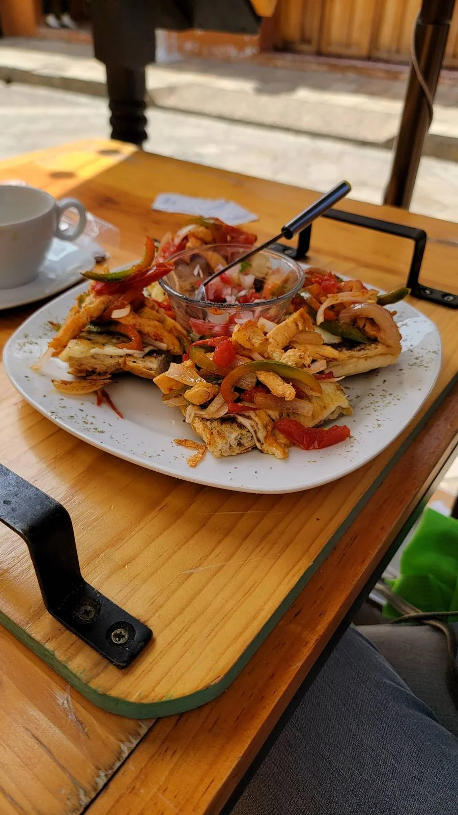 A plate of grilled fish topped with sautéed vegetables, including onions and red bell peppers, served on a wooden table outdoors. There's an empty white cup and saucer beside the plate.