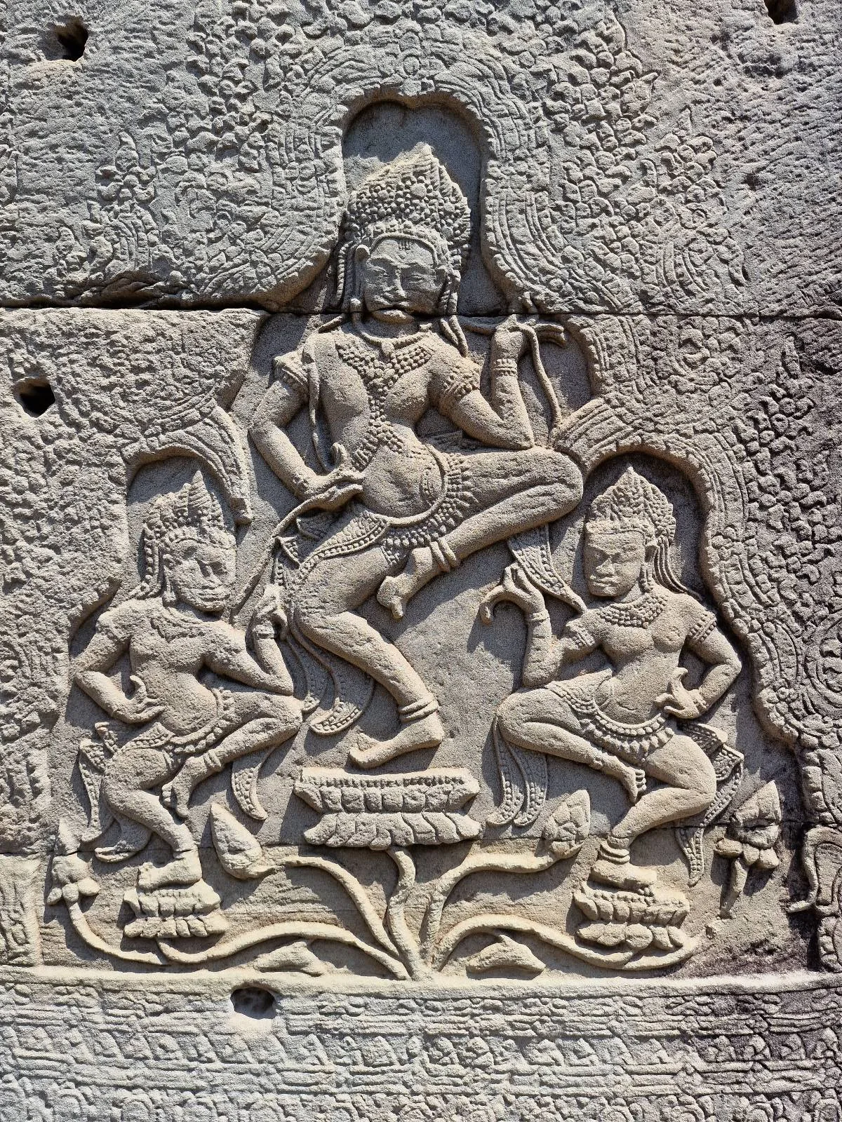 Stone carving depicting a central dancing figure with two attendants on either side, surrounded by ornate patterns, likely part of a temple or ancient structure.