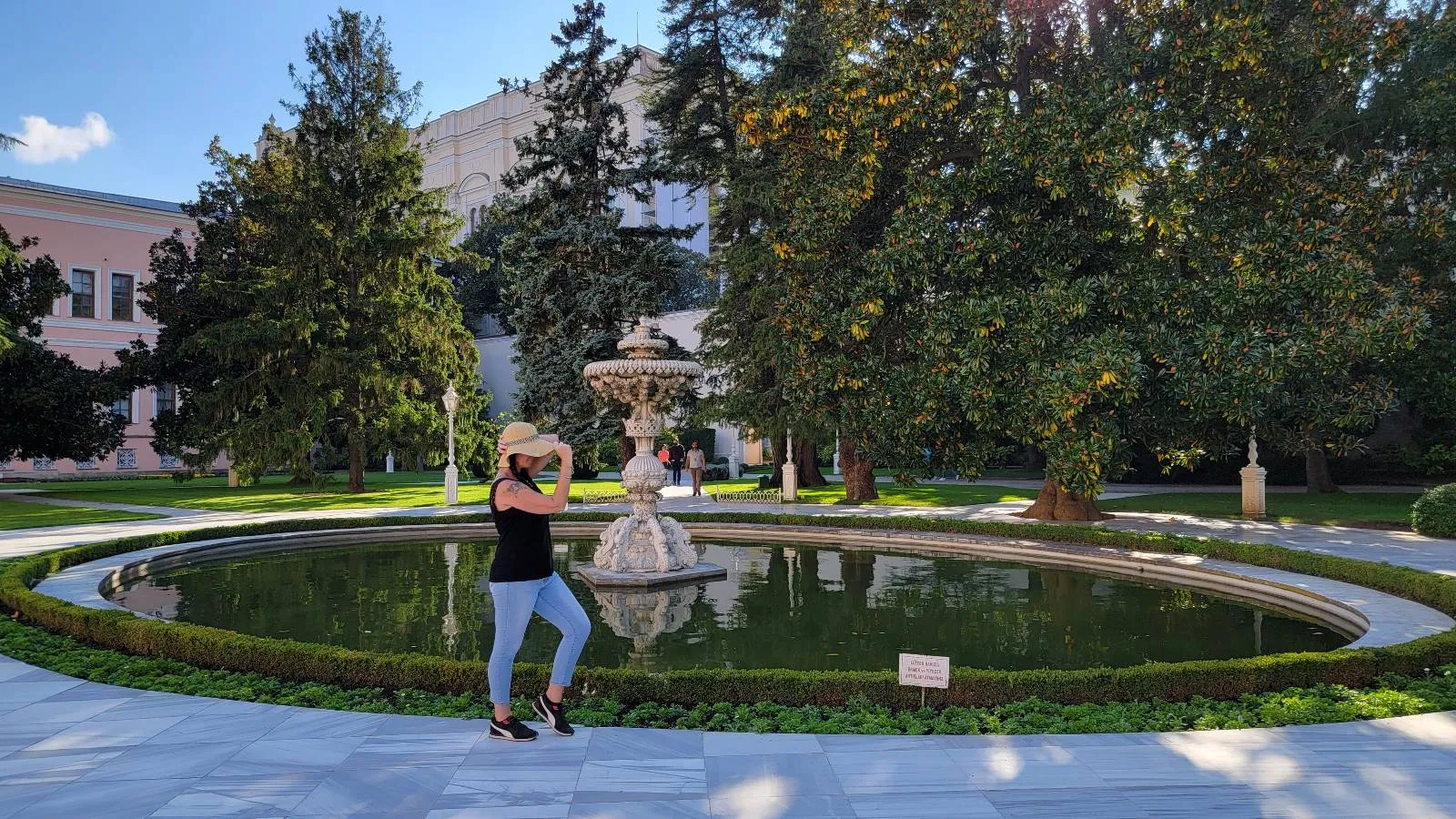 A woman in a hat poses for a photo beside a circular fountain in a garden, surrounded by green trees and a building in the background on a sunny day.