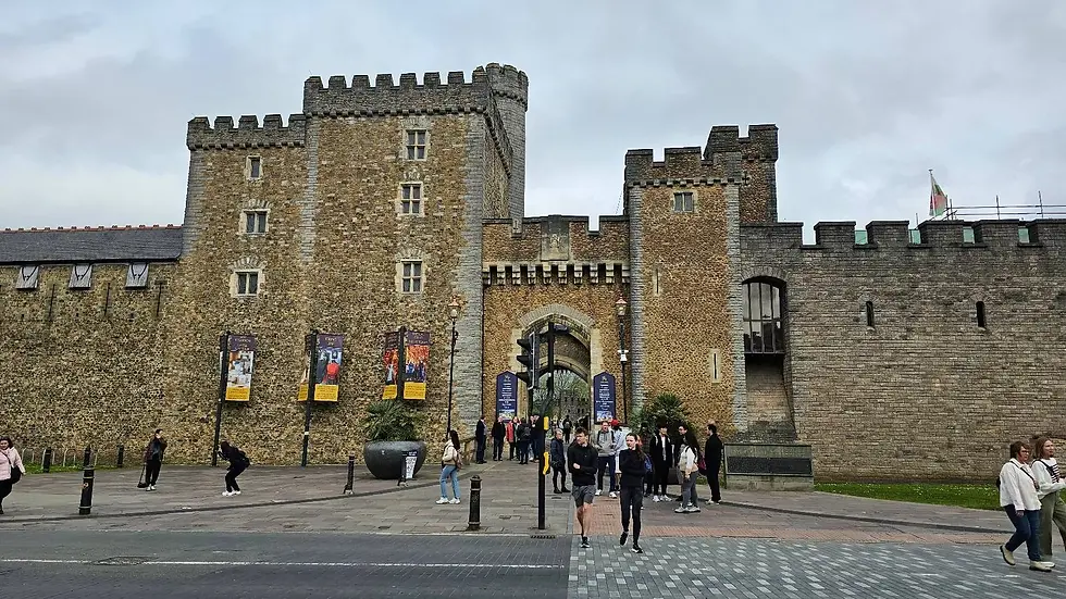 A historical stone castle with a large arched entrance and banners, with people walking and interacting in front.