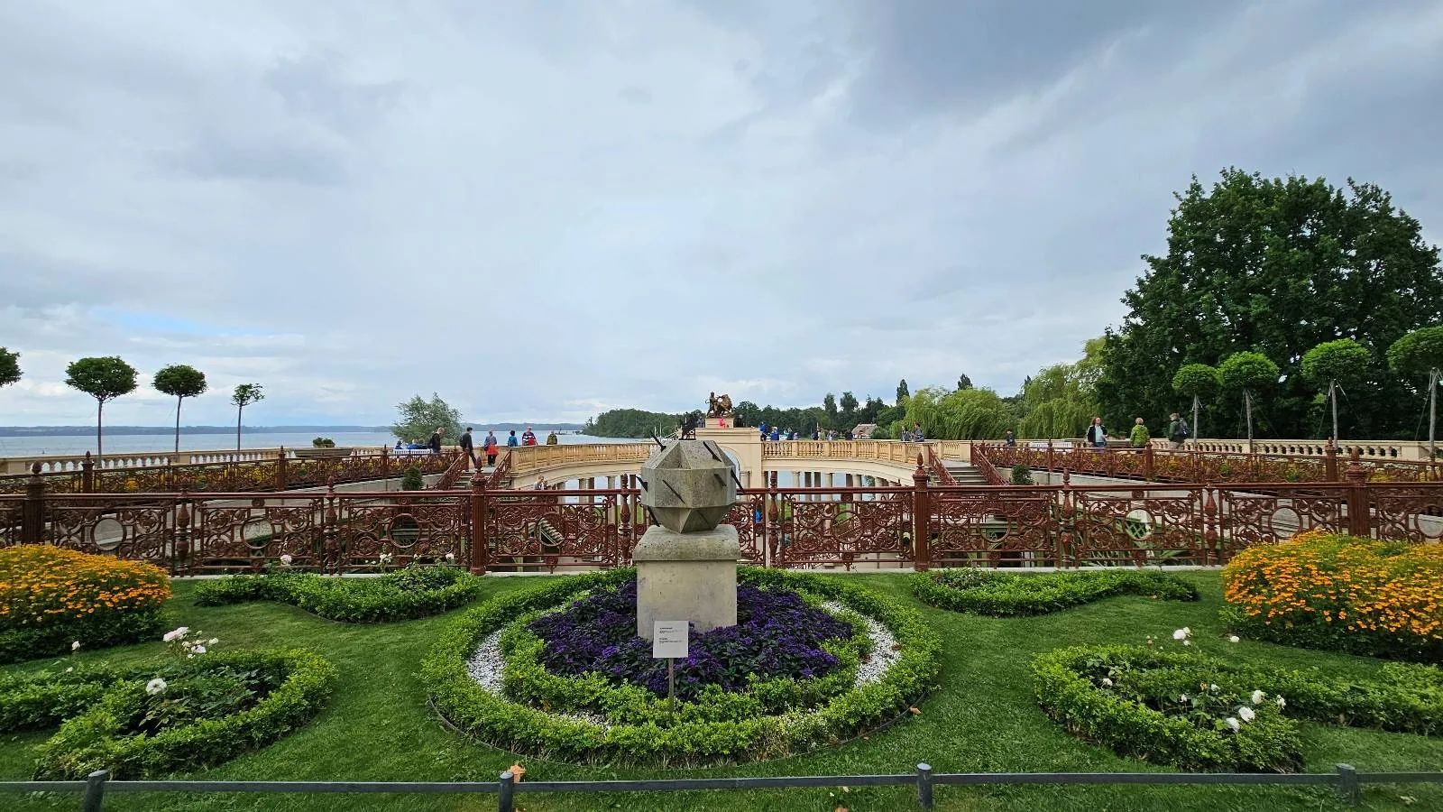 A landscaped garden with neatly trimmed bushes, colorful flower beds, and a stone statue in the center, overlooking a sandy beach and a large body of water under a cloudy sky.
