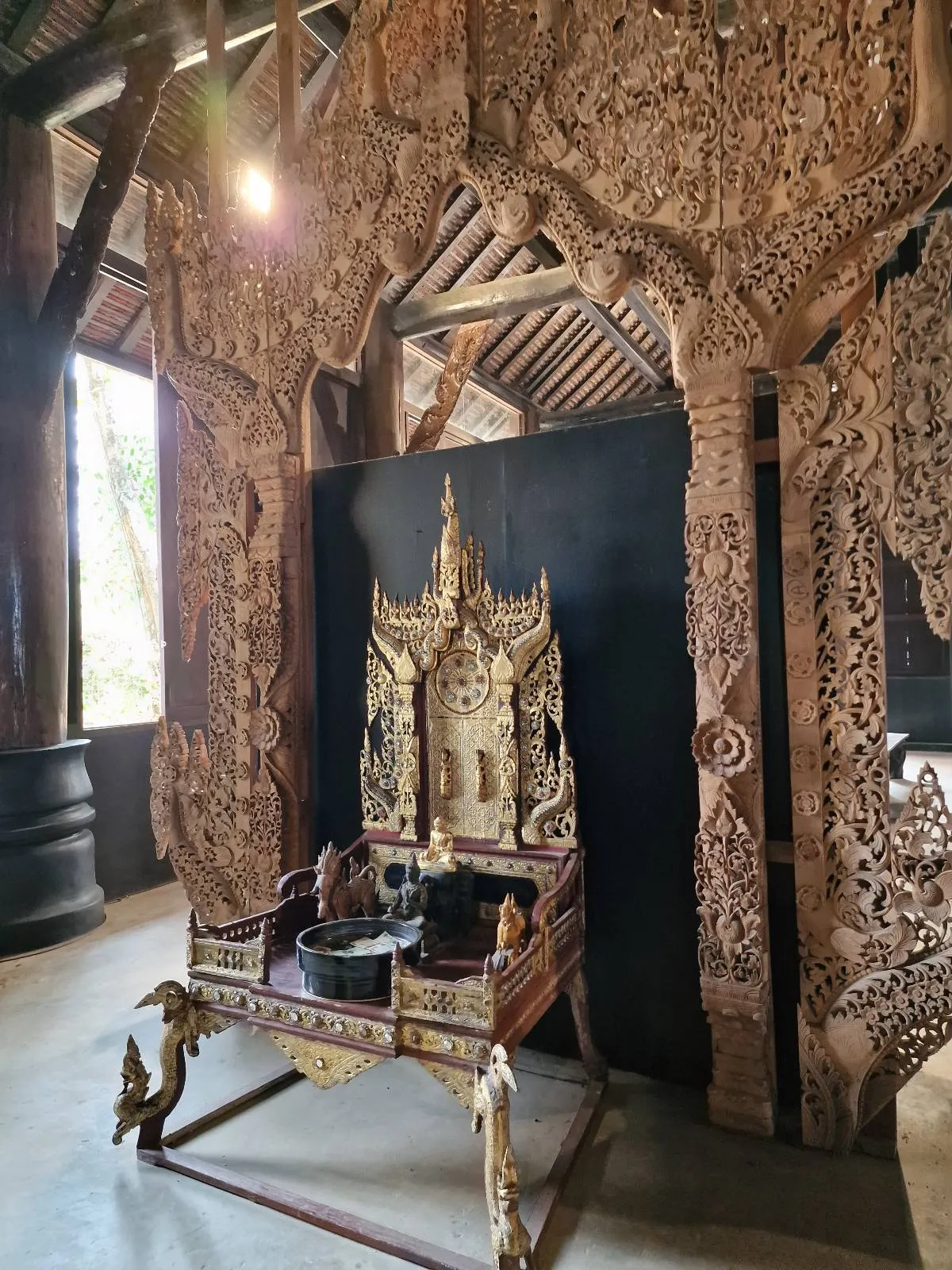 Ornate carved wooden structure with intricate designs, featuring a decorative chair and small sculptures, set against a dark backdrop in a well-lit room.