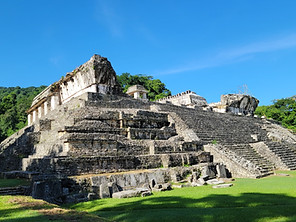 Ancient stone pyramid with stairs and platforms, surrounded by lush greenery under a clear blue sky, reminiscent of ancient ruins in Mexico. The structure has weathered surfaces and a small tree nearby.