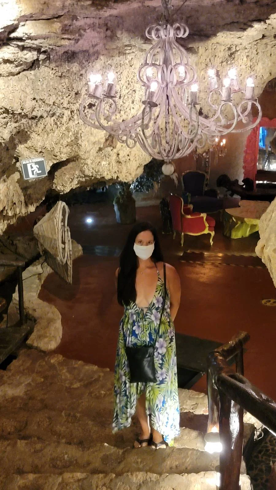A woman wearing a floral dress and face mask stands on stone stairs in an underground cave-themed restaurant. A large chandelier hangs above, illuminating rustic furniture and decor.
