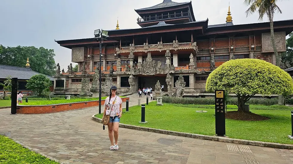 Woman in casual attire stands on a stone path in front of a decorative building, with lush landscaping and a cloudy sky in the background.