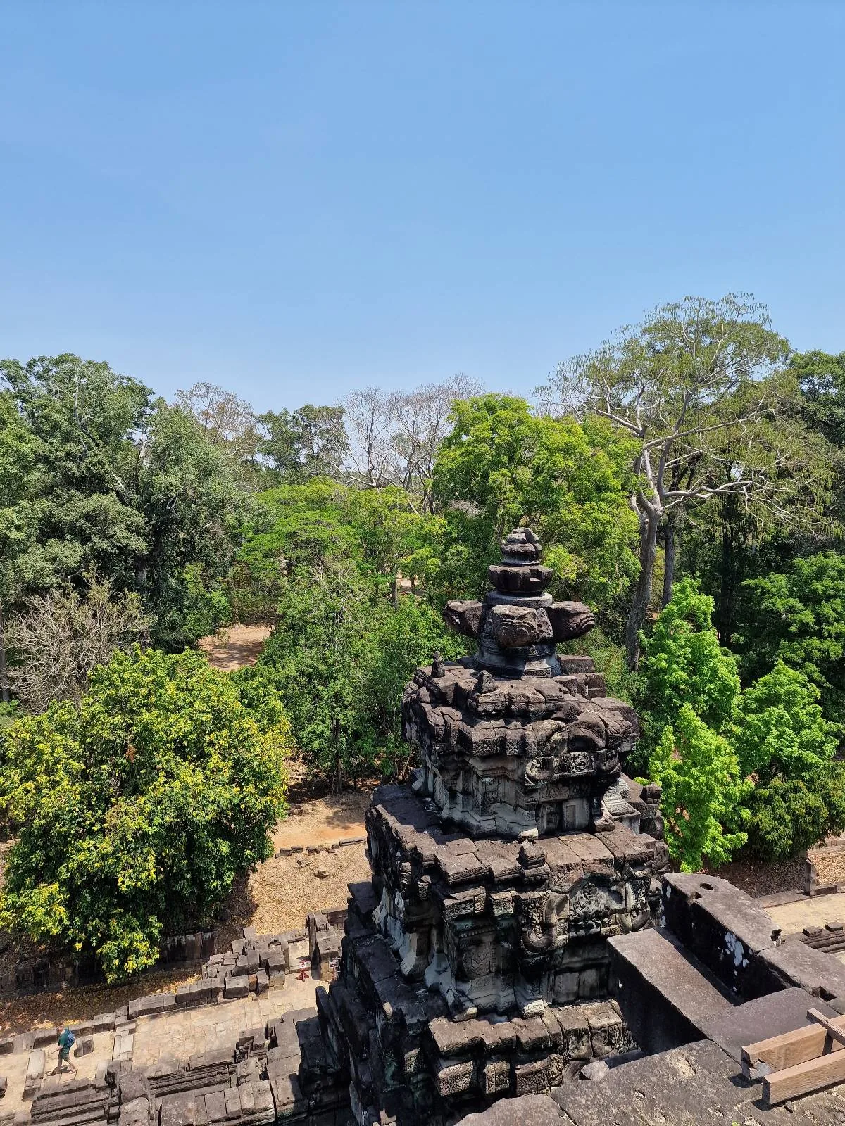 Ancient stone temple with tiered roof surrounded by lush green trees under a clear blue sky, viewed from above.