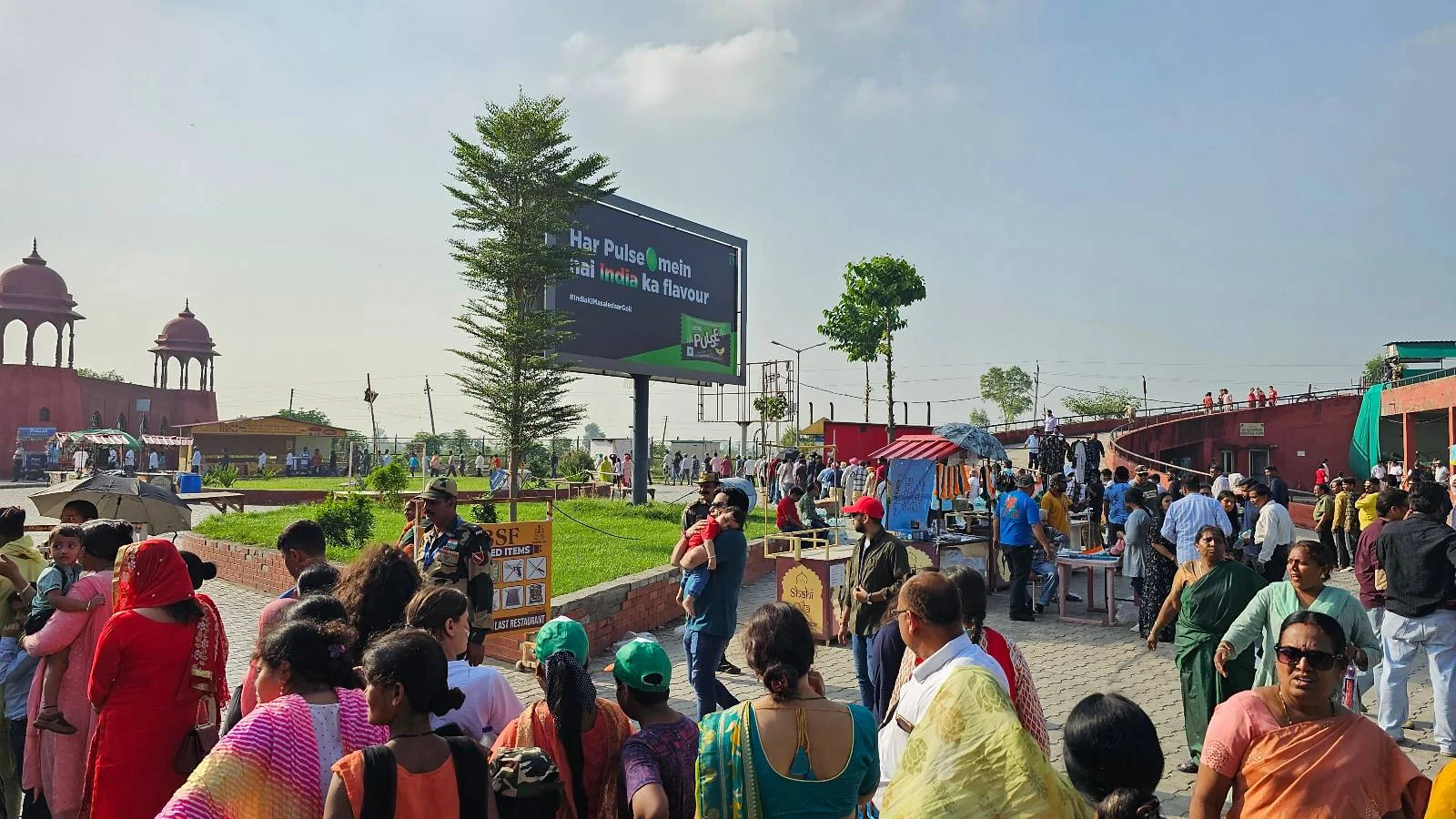 A large crowd gathers near the entrance of a historic site with red buildings and domes, lush green lawns, and a big digital signboard under a clear blue sky. People are walking, talking, and taking photographs.