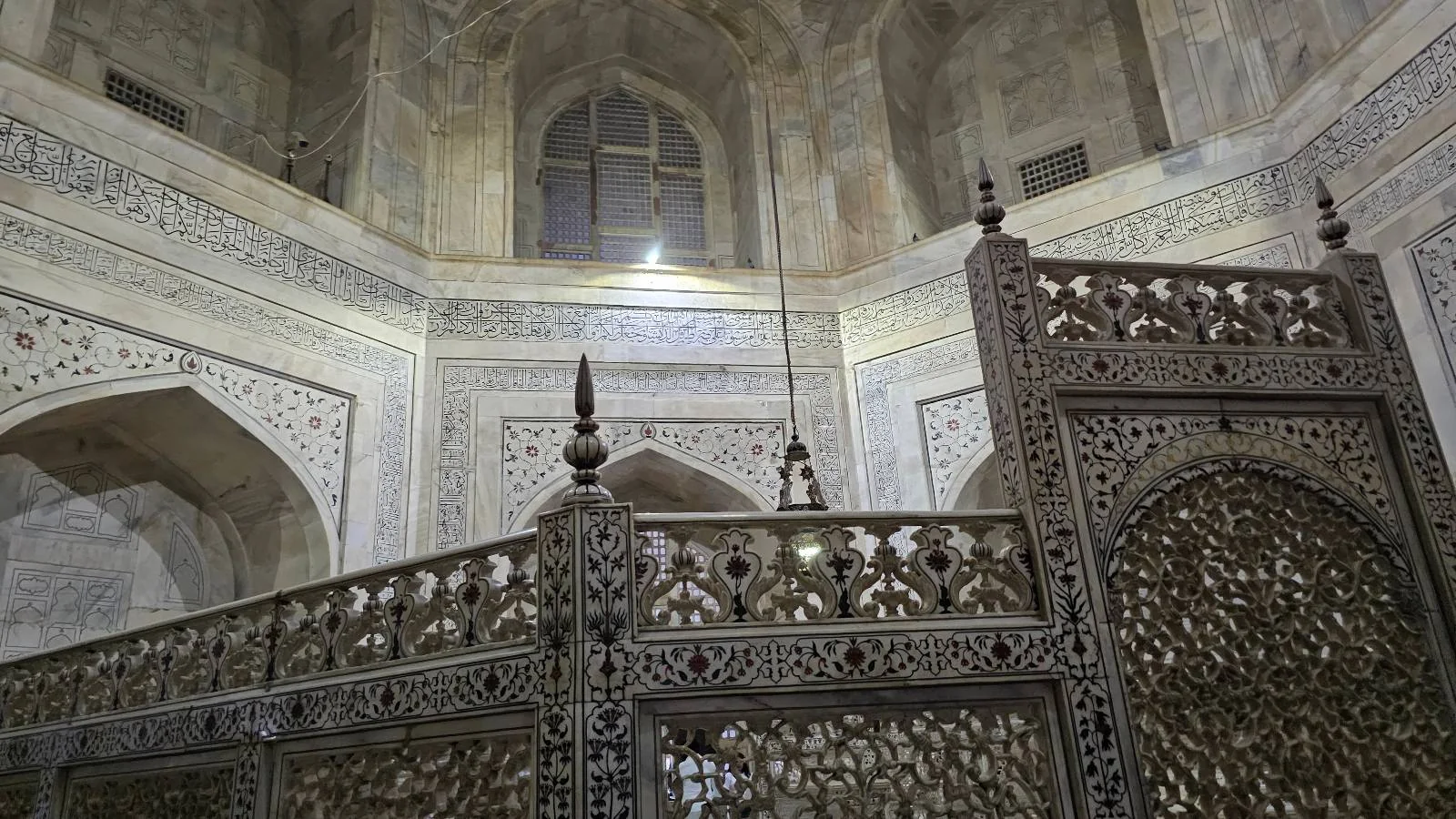 Intricately carved stone or metal lattice screen in an old building, with arches, a window above, and ornate architectural details on the walls in soft lighting.
