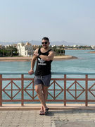 A man in a black tank top and shorts leans against a railing, pointing towards the water in El Gouna, with its beautiful beaches framed by a backdrop of houses, mountains, and a clear sky.