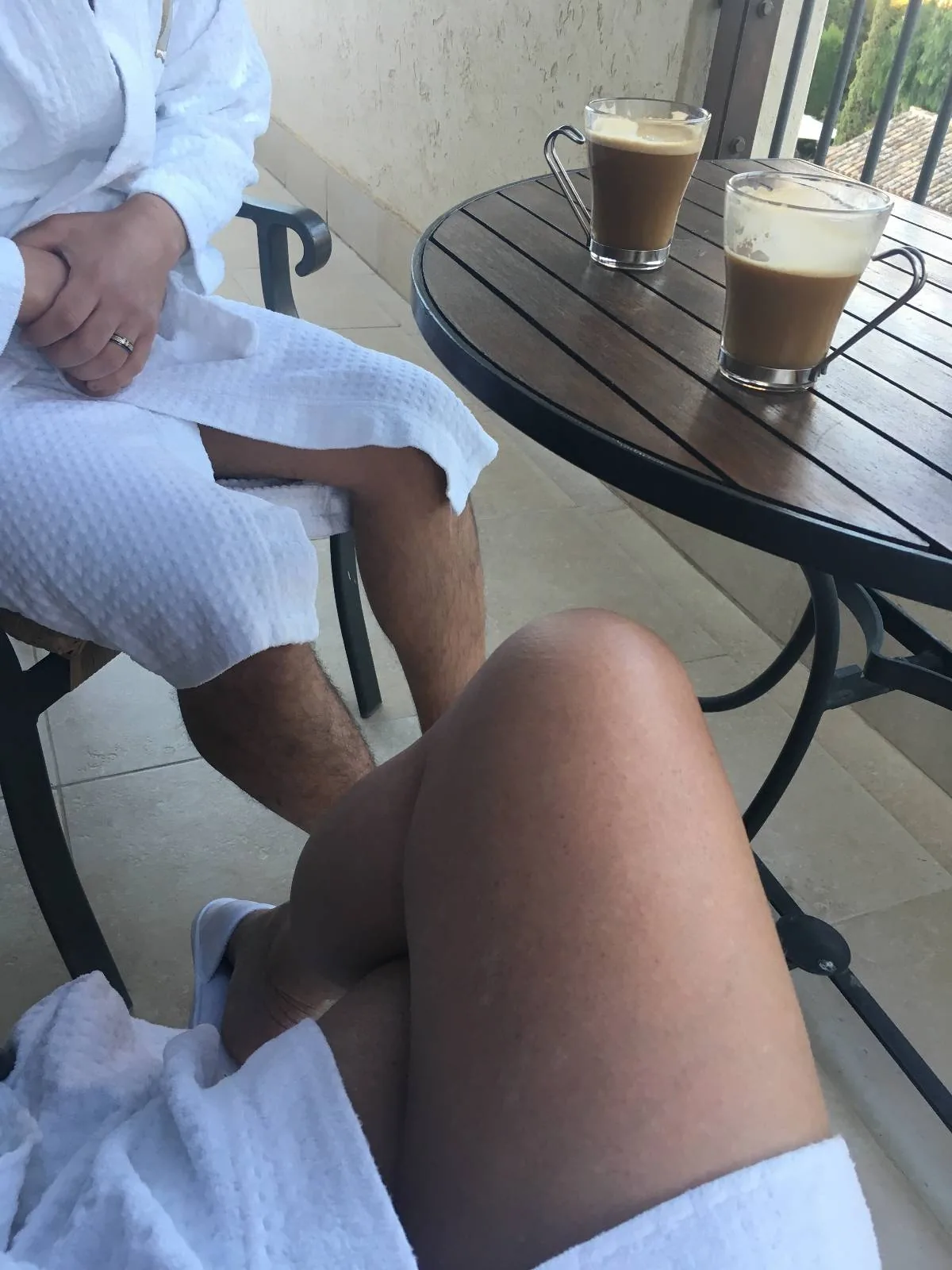 Two people in white robes are sitting at a round wooden table. Two cups of frothy coffee are on the table. They are sitting outside on a patio with a railing in the background.