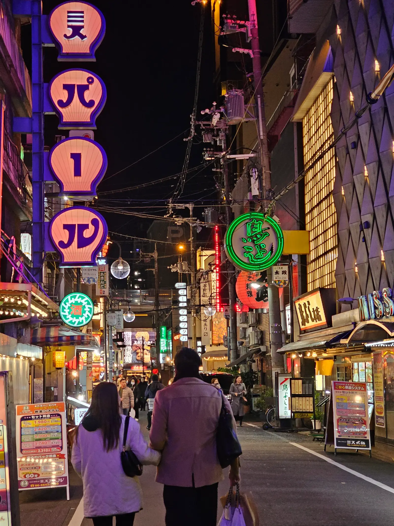A couple walks down a lively city street at night, surrounded by illuminated signs in various colors and scripts, with restaurants and shops lining both sides of the street.