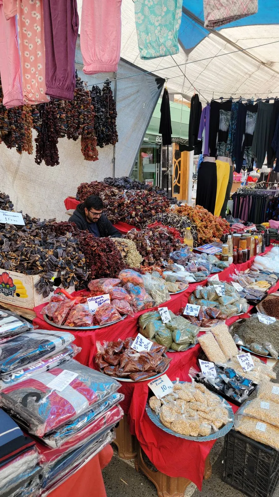 Market stall with a variety of dried fruits, nuts, and textiles on display. A vendor is sitting behind the items under a canopy.