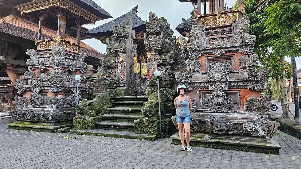 Person in helmet stands by ornate Balinese temple with intricate carvings. Stone statues and greenery surround, creating a serene atmosphere.