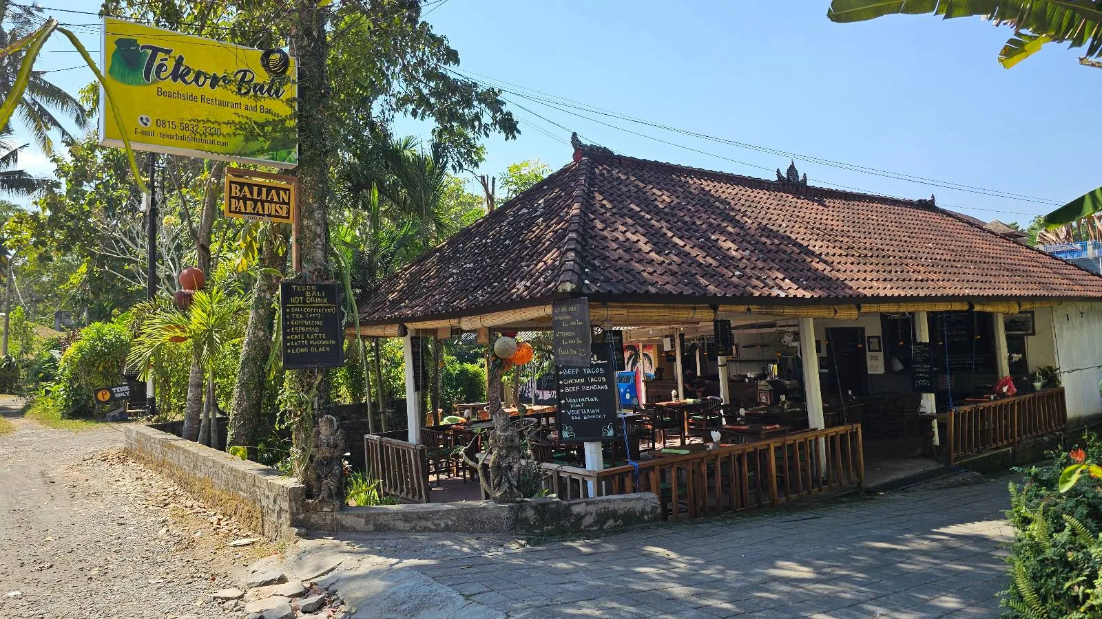 A quaint restaurant with a thatched roof and open-air seating surrounded by lush greenery. A signpost at the entrance displays the restaurant's name and menu offerings. The bright sunny day highlights the tropical ambiance.