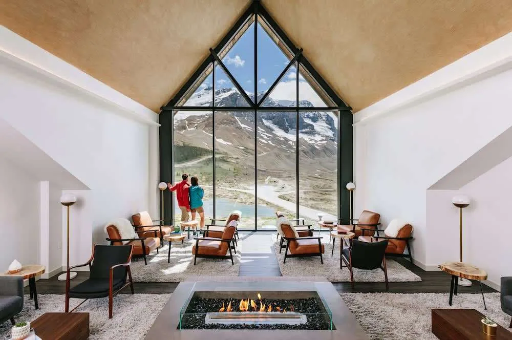 A spacious lounge with tall triangular windows frames a snowy mountain view. Comfortable chairs surround a modern fireplace. Two people stand by the window, one in red and the other in blue, admiring the scenery.