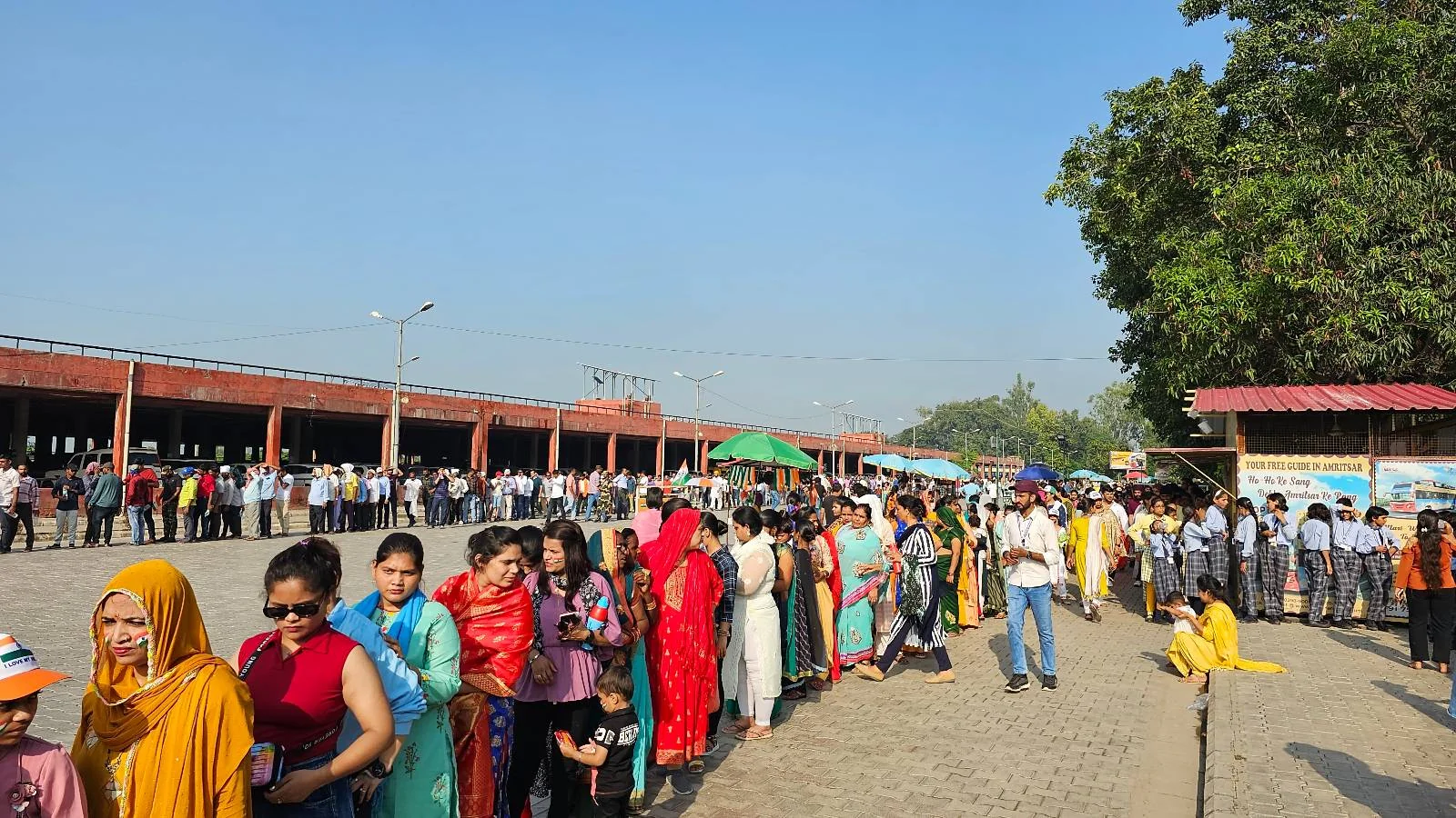 A large group of people, including women in colorful clothing, stand in a long queue outdoors under clear blue skies, with some trees and a building visible in the background.