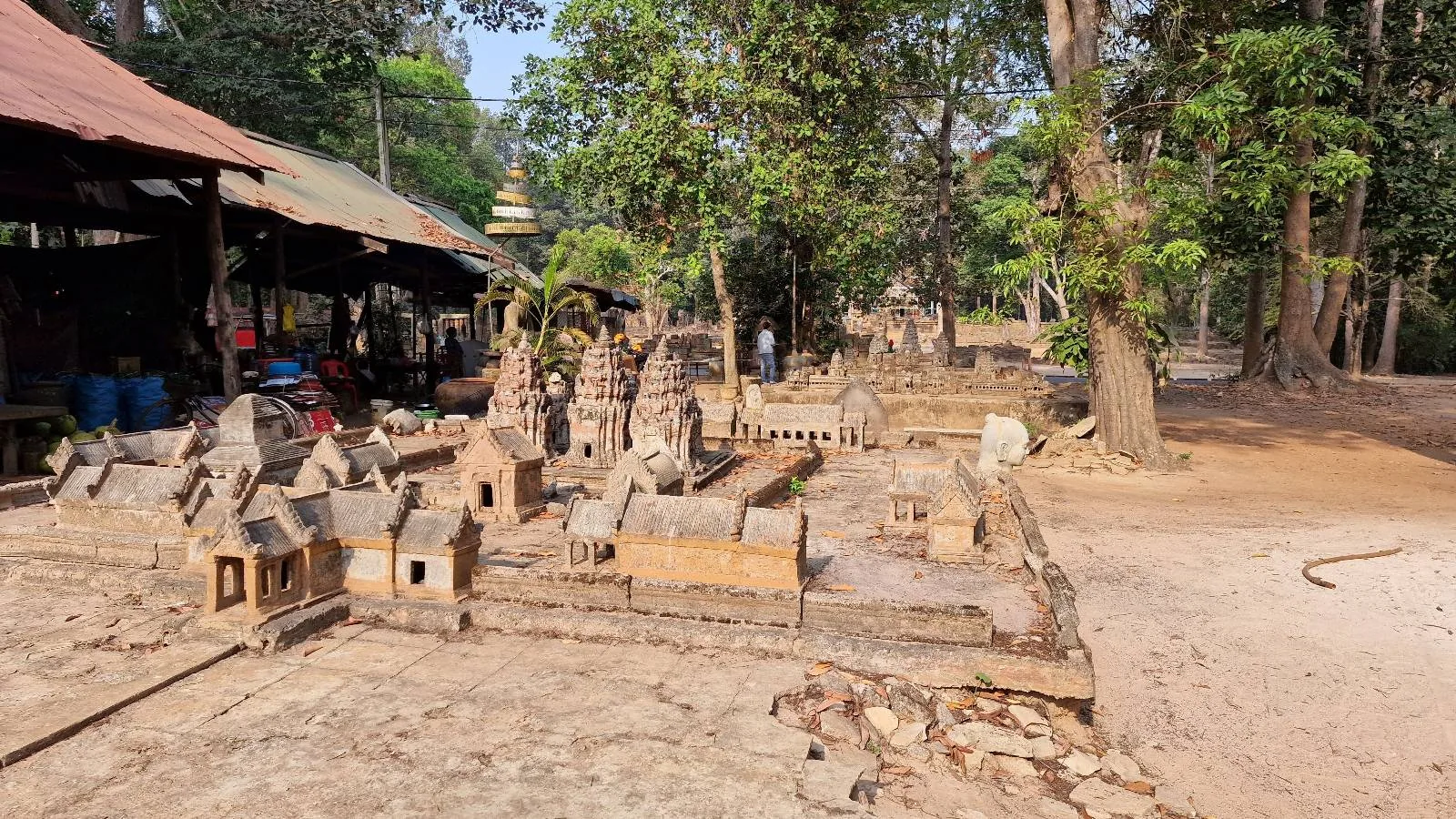 A miniature model of an ancient temple complex surrounded by trees. The structures are made of stone and are set outdoors under a clear sky. Some wooden huts and greenery can be seen in the background.