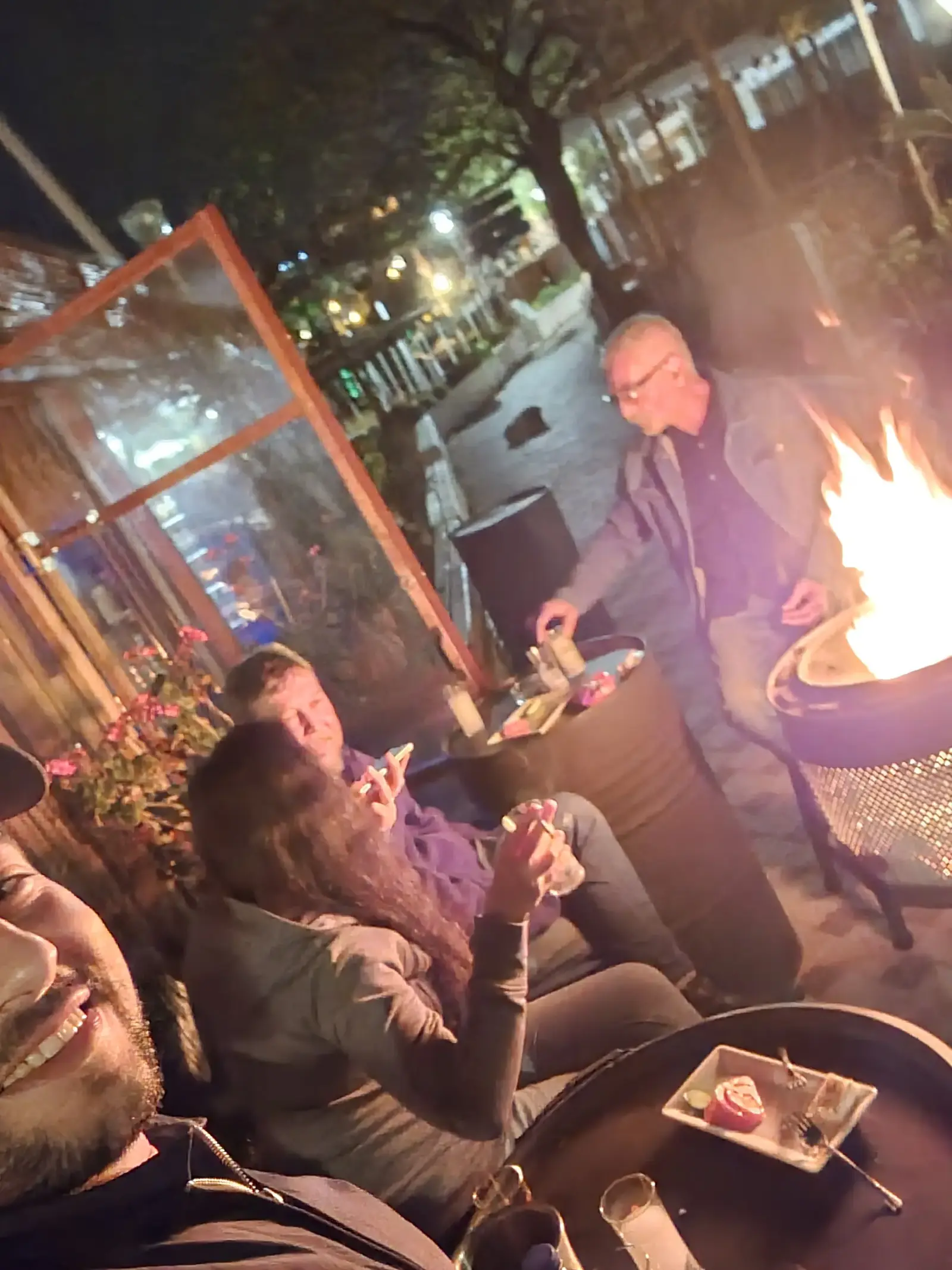 A group of people sitting around a fire pit at night, chatting and eating outdoors.