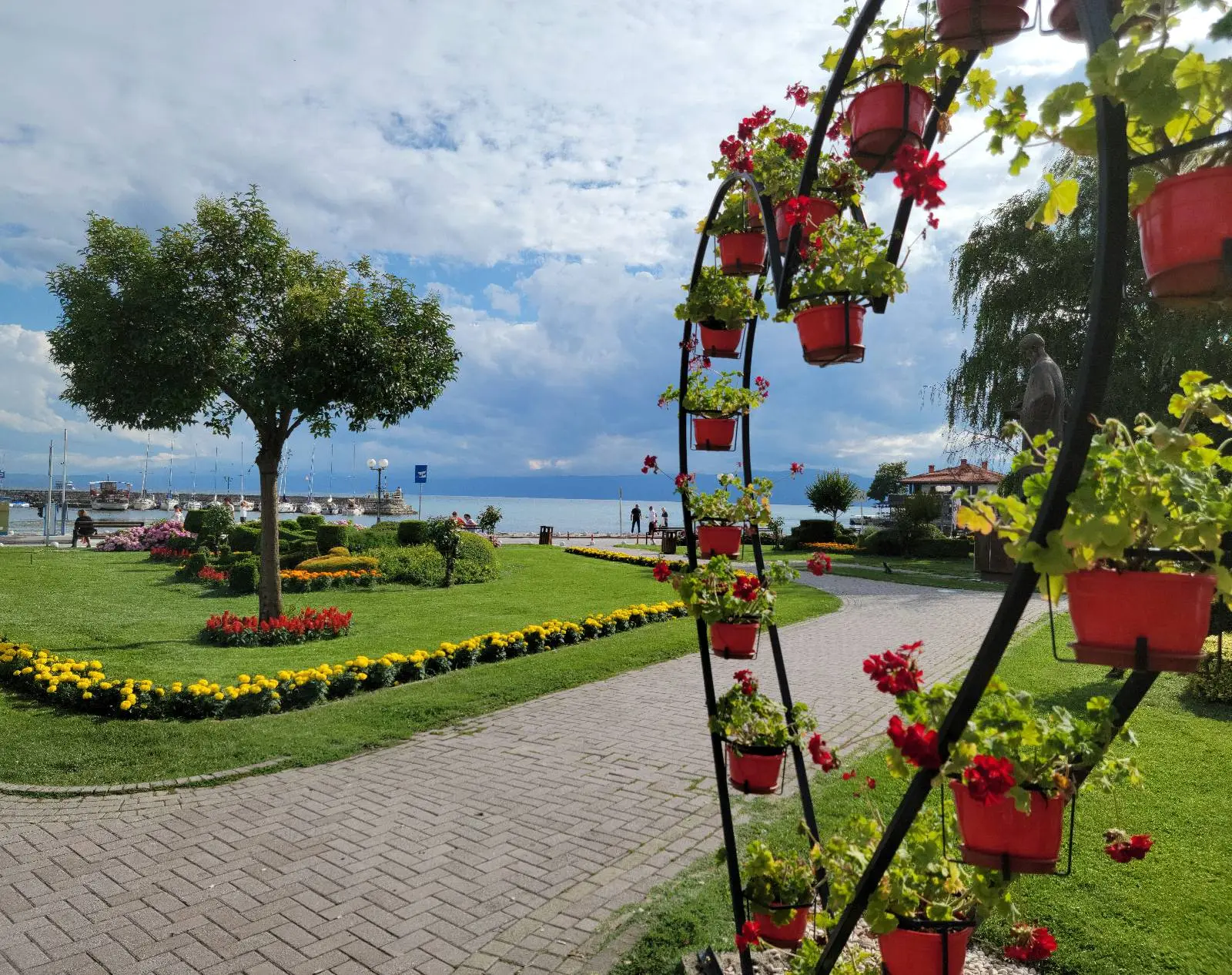 A scenic lakeside park with a winding brick path lined with vibrant red and yellow flowers. Red flower pots hang on a decorative metal arch. A large tree provides shade, and the lake and sky are visible in the background.