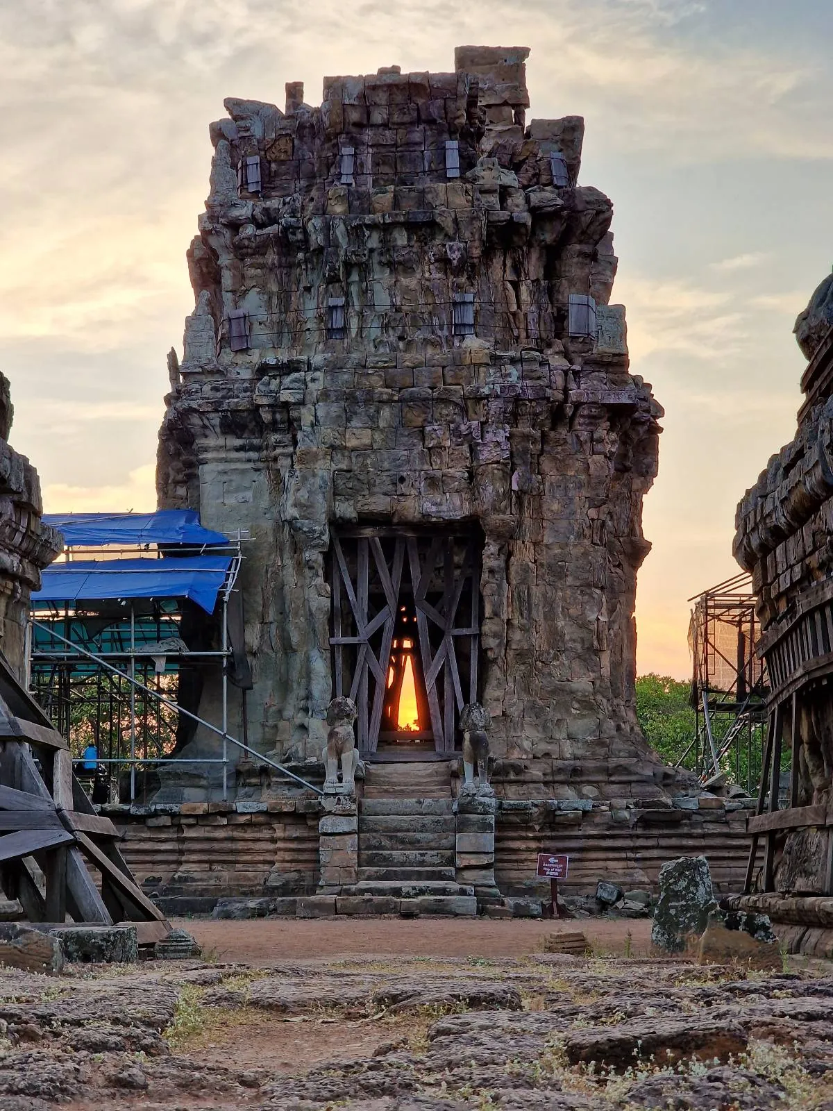 Ancient stone temple with a central tower partially restored, wooden supports at the entrance, and a warm light glowing inside at sunset. Ruins and scaffolding are visible, with trees and sky in the background.