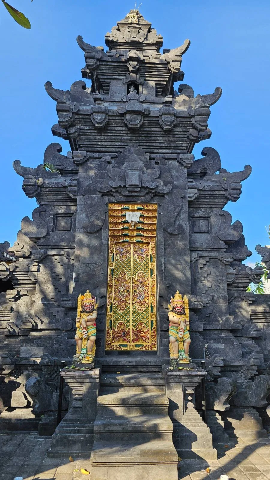 Ornate stone temple gate with intricate carvings and a decorative wooden door. The gate is adorned with statues of figures on either side. The sky is clear and blue, casting shadows on the structure.