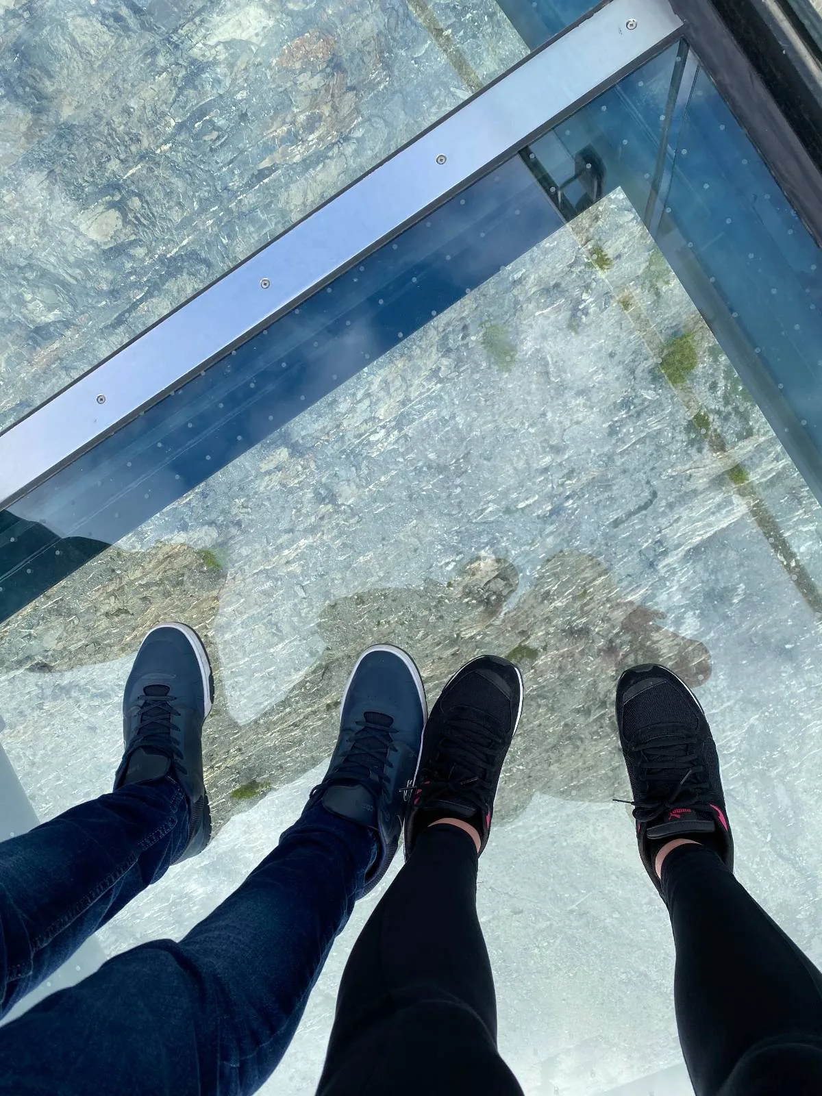 Two people stand on a transparent glass floor, looking down at a rocky landscape far below. Only their legs and shoes are visible in the photo. The glass creates a sense of height and openness.