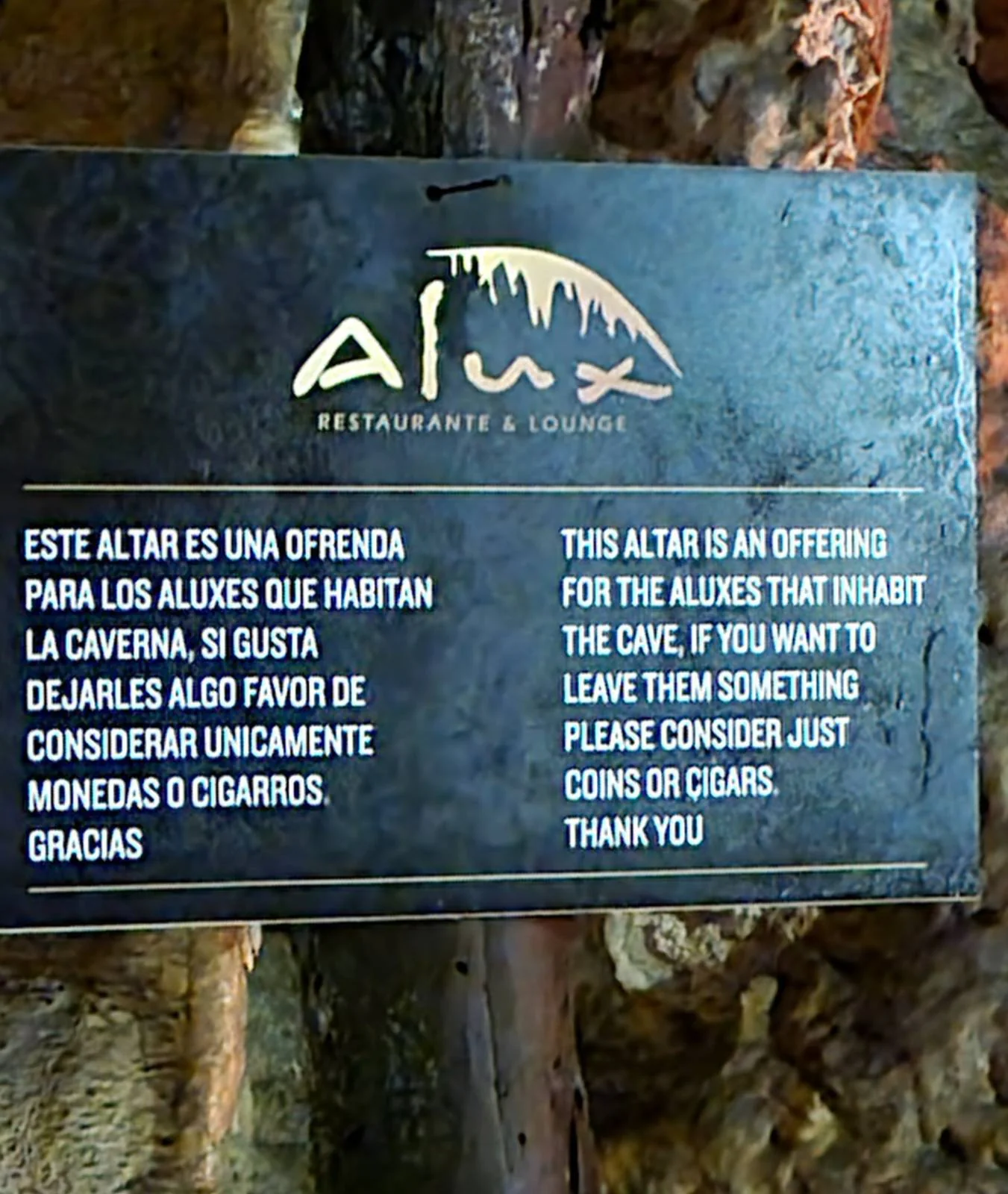 Sign with text in Spanish and English. Spanish text: invitation to leave offerings for the aluxes in the cave. English text: "This altar is an offering for the aluxes that inhabit the cave. If you want to leave them something, please consider just coins or cigars. Thank you.