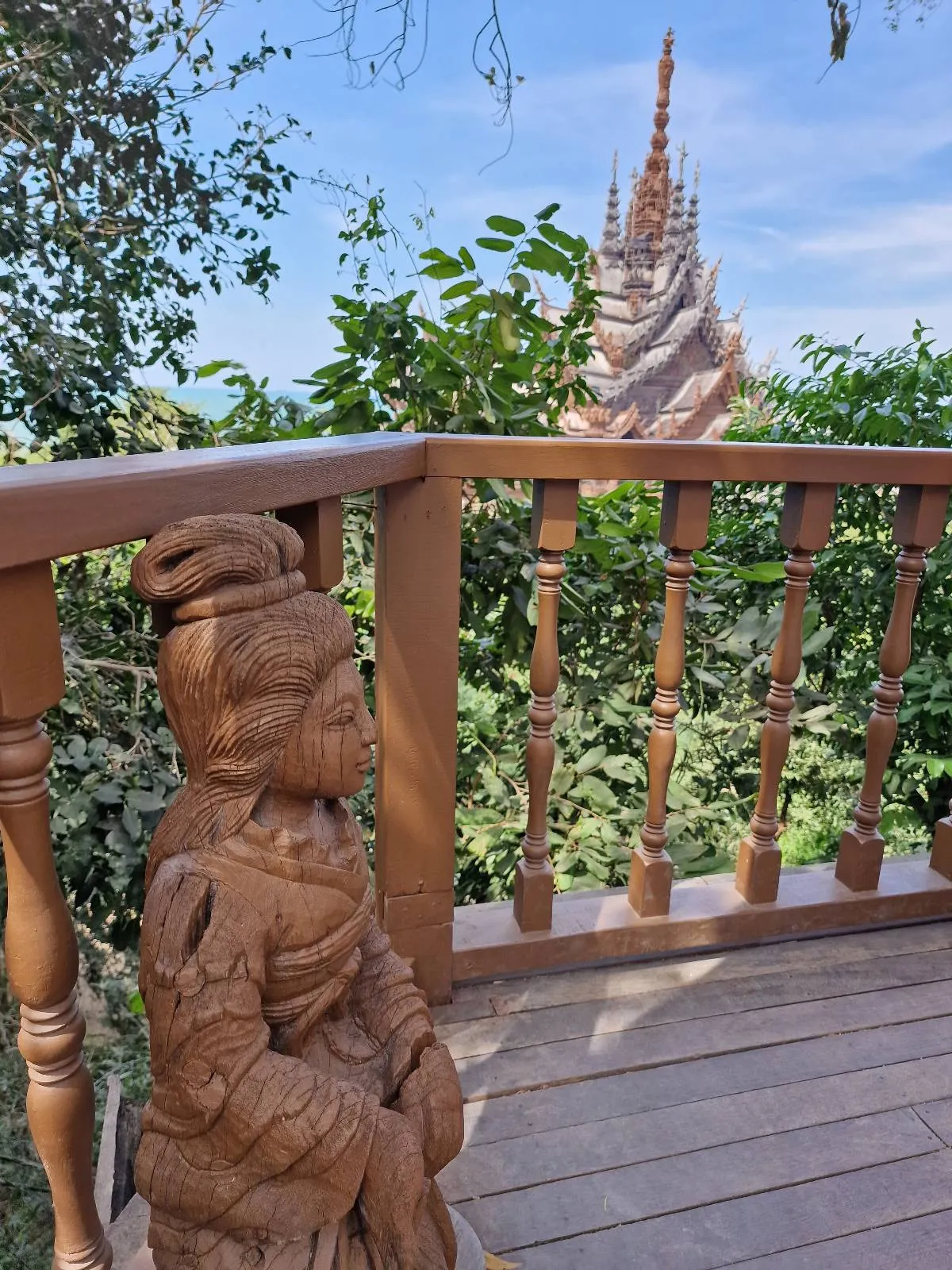 A wooden carved statue stands on a deck with a wooden railing, overlooking lush green foliage and a pointed temple structure in the background under a blue sky.