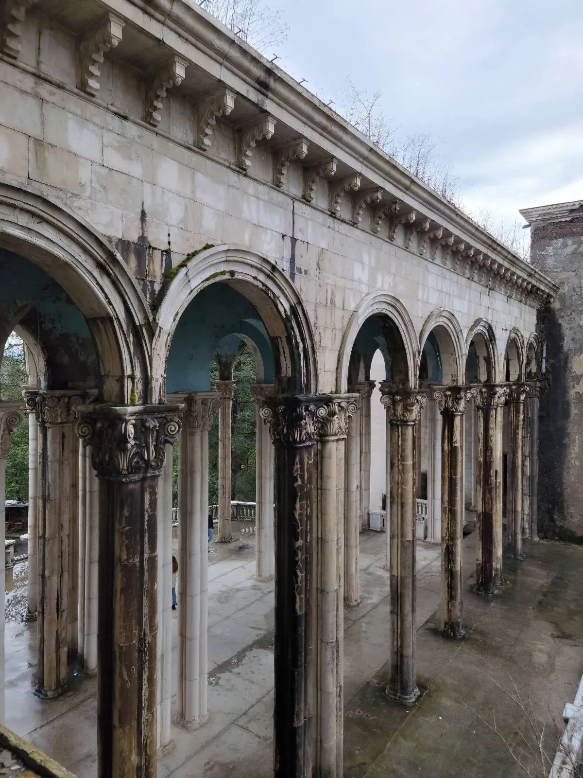 Stone arches of a historic ruined building with tall columns, partially open to the sky, and surrounded by greenery, under a cloudy sky.