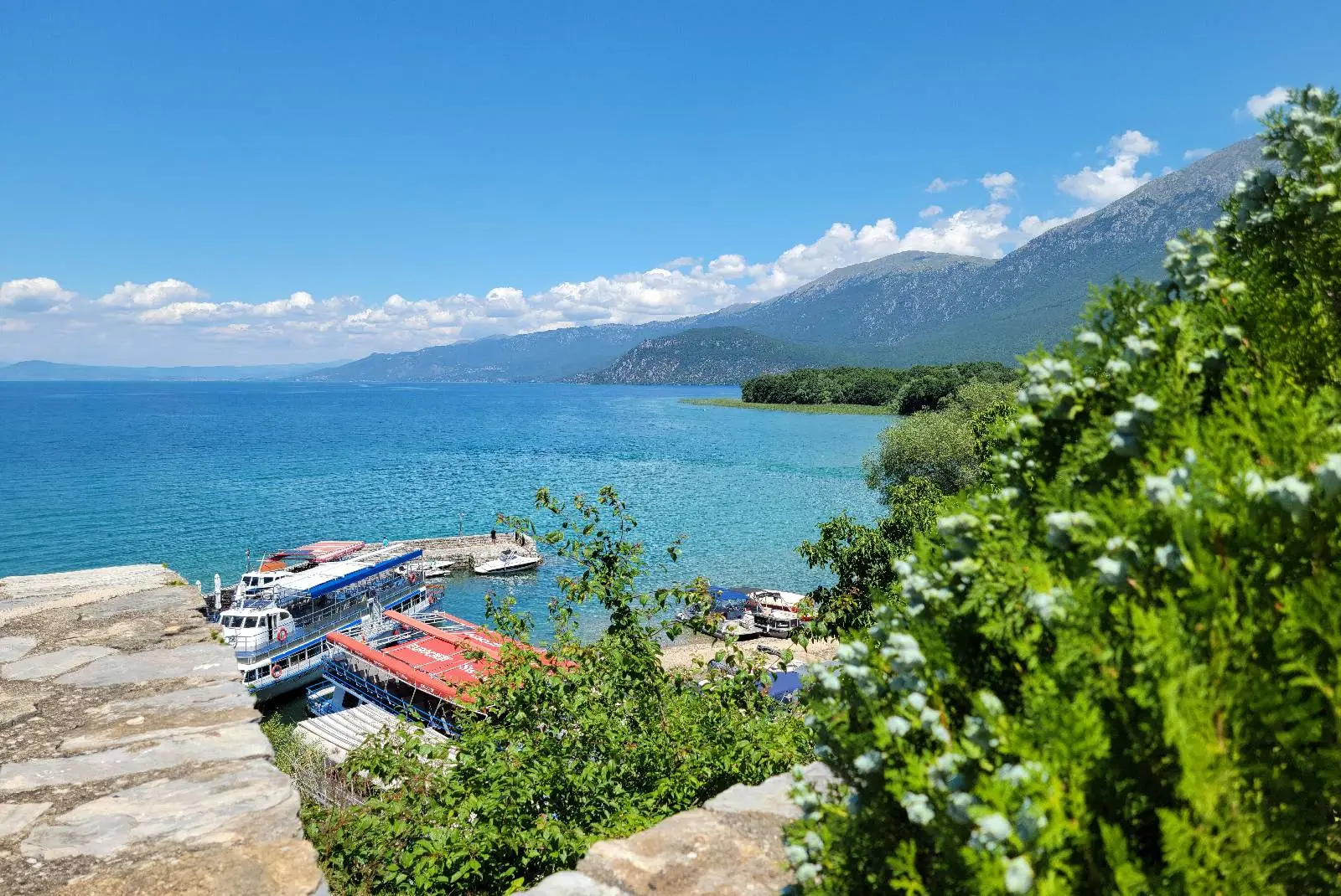 Scenic view of a clear blue lake with several boats docked by a stone pier. Lush greenery and flowering bushes are in the foreground. Mountains under a bright blue sky with scattered clouds form the background.