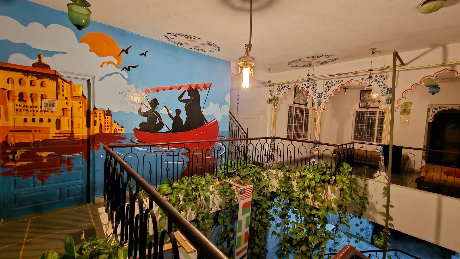 A brightly colored wall mural shows silhouettes of two people rowing a boat at sunset, with an orange palace and birds in the background. The indoor space features green plants and hanging lights.